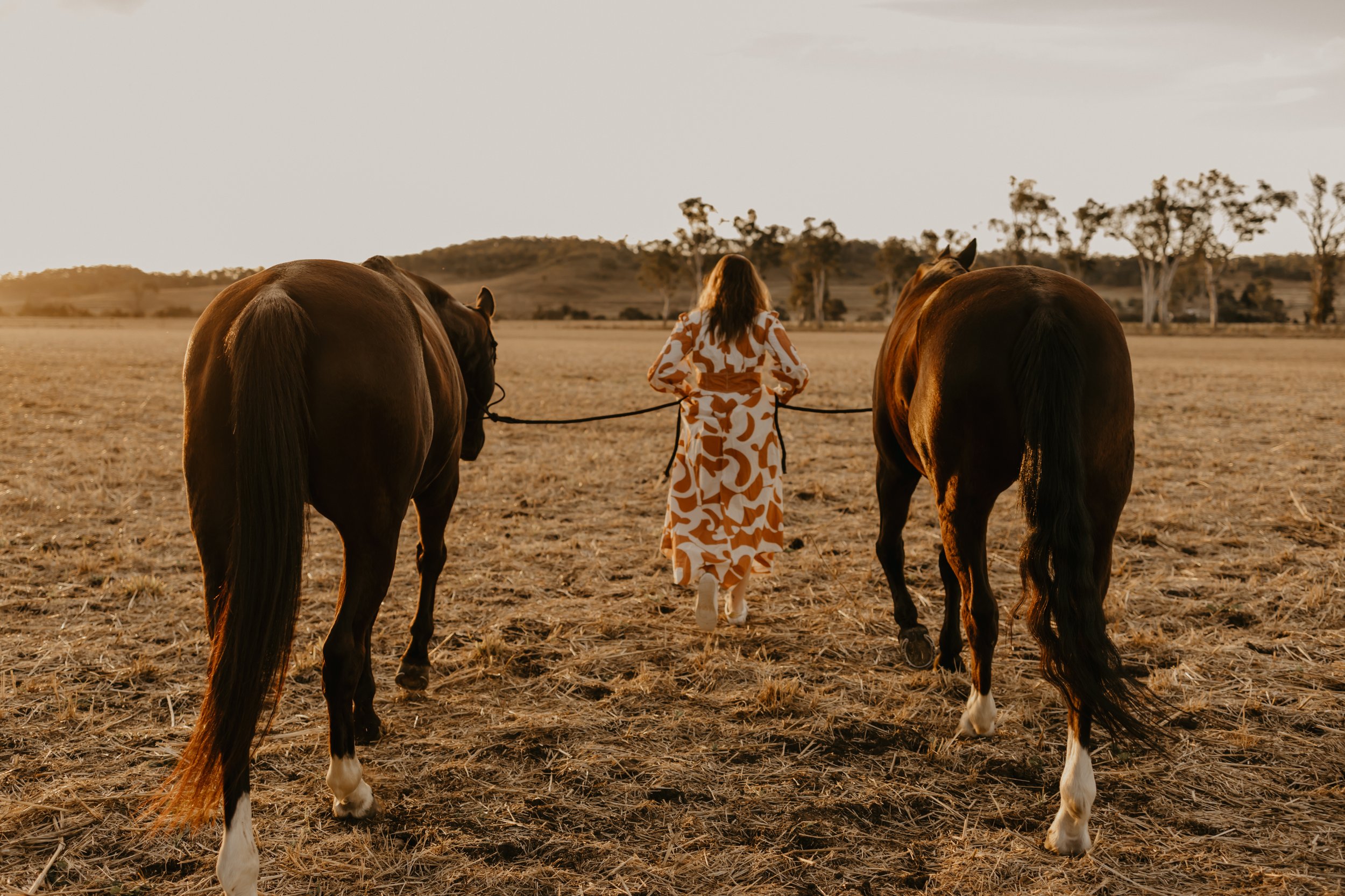 Woman in a patterned dress walking with two horses in an open field during sunset.