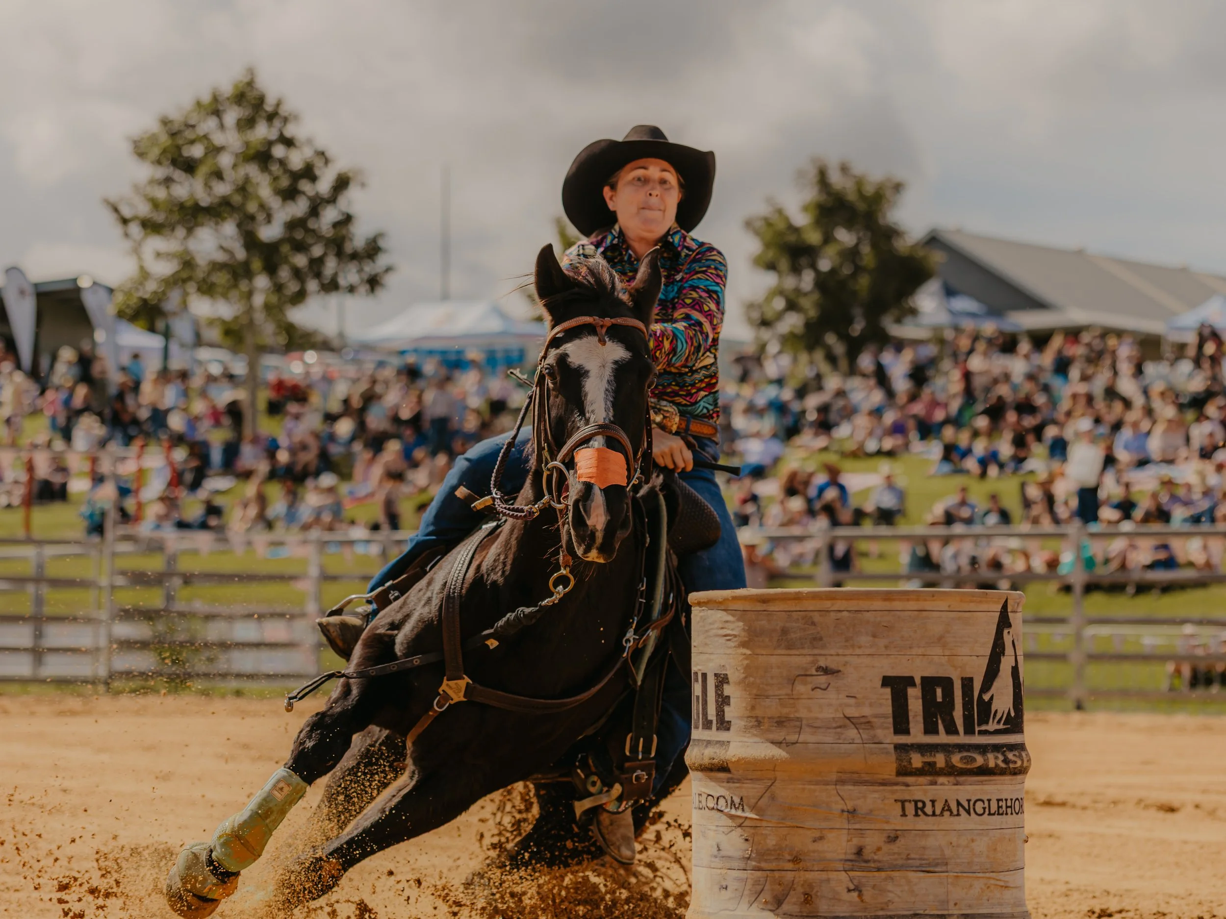 The WESTRN Edit | Western Sports Photography - A person wearing a colorful shirt and a cowboy hat is riding a horse and jumping over a wooden obstacle, with a crowd watching in the background at an outdoor rodeo or equestrian event.