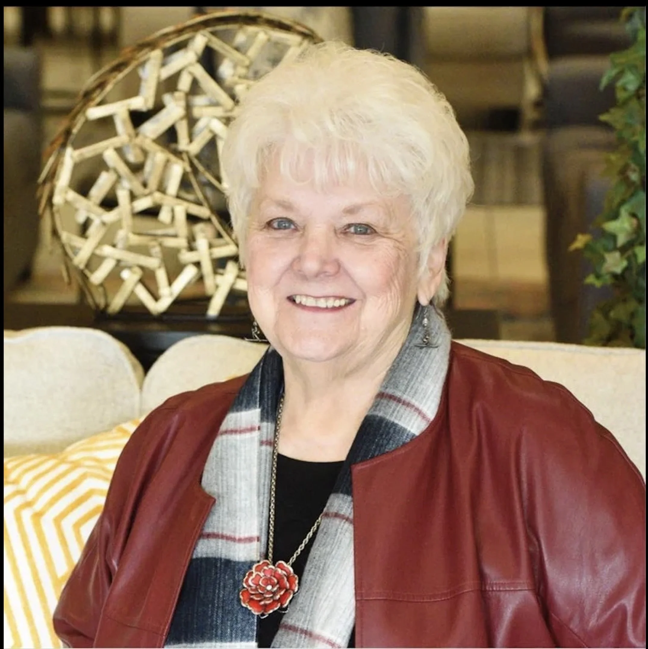 Close-up of an elderly woman with short, curly white hair, wearing a red leather jacket and a large red flower necklace, standing in front of a decorative wooden star sculpture.