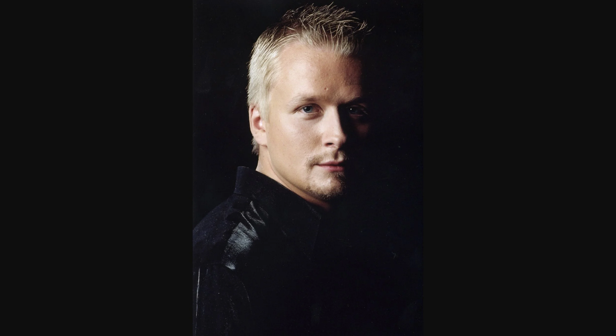 Close-up of a young man with blond hair and blue eyes, wearing a black velvet jacket, posing against a dark background.