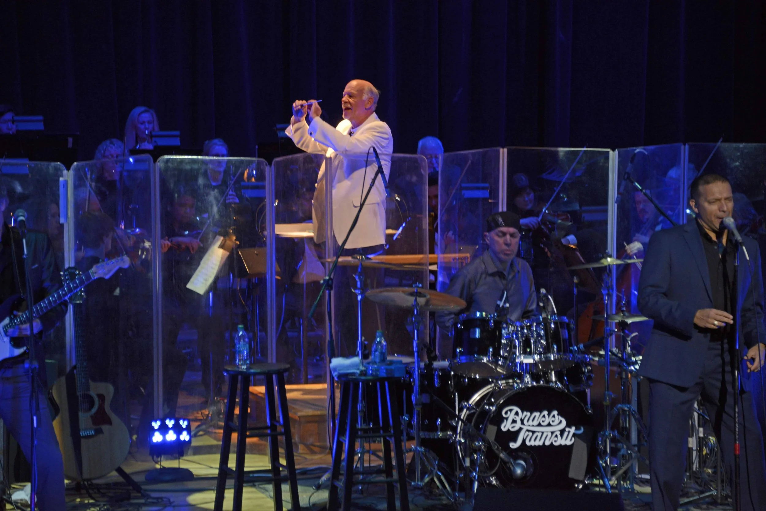 An elderly man in a white suit conducting a live band on stage, with musicians playing guitars, drums, and keyboards behind transparent screens, and music stands, in a dark theater setting.
