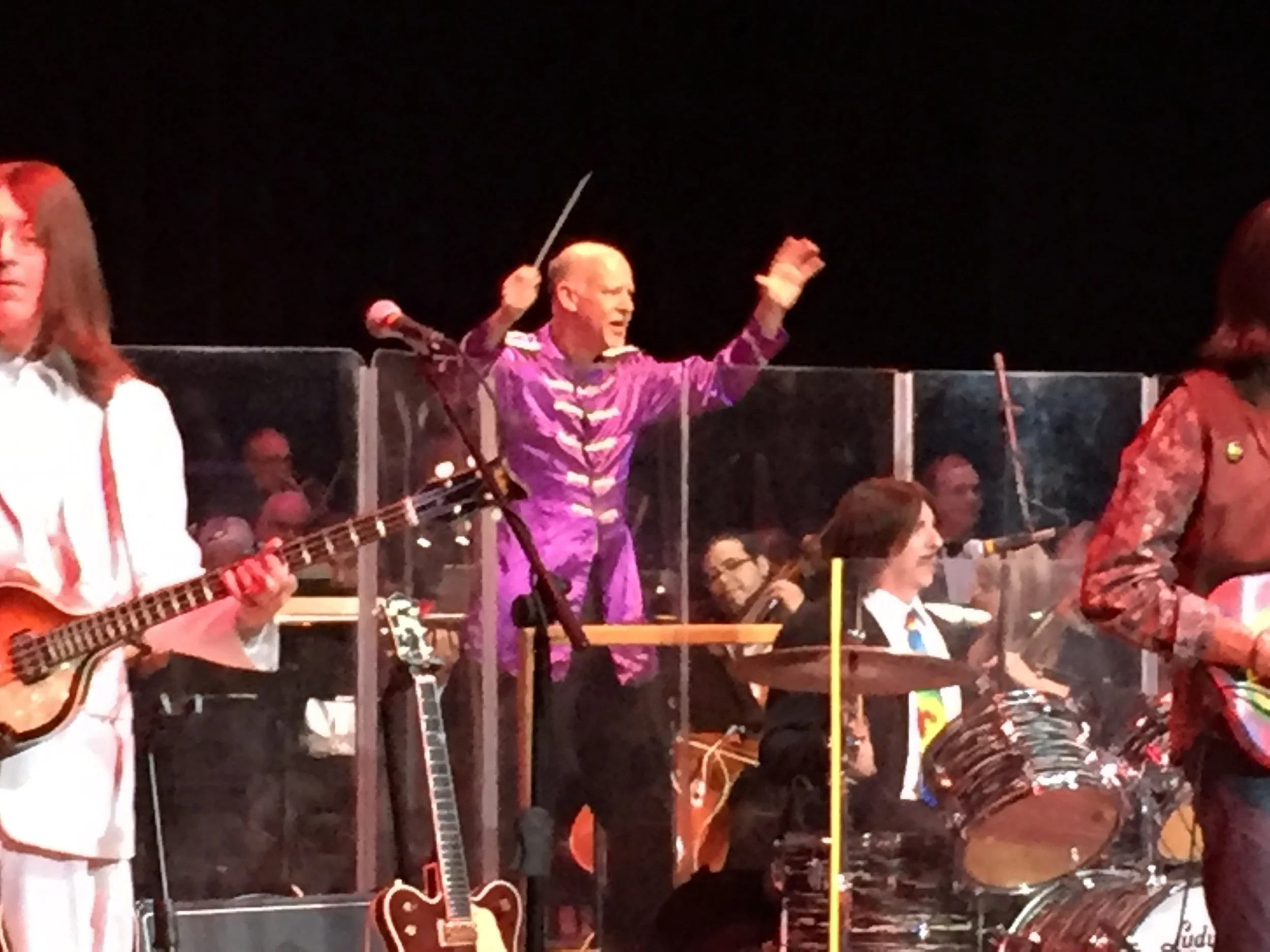 A group of musicians, including a person conducting, performing on stage at night with musical instruments and people in the audience visible.