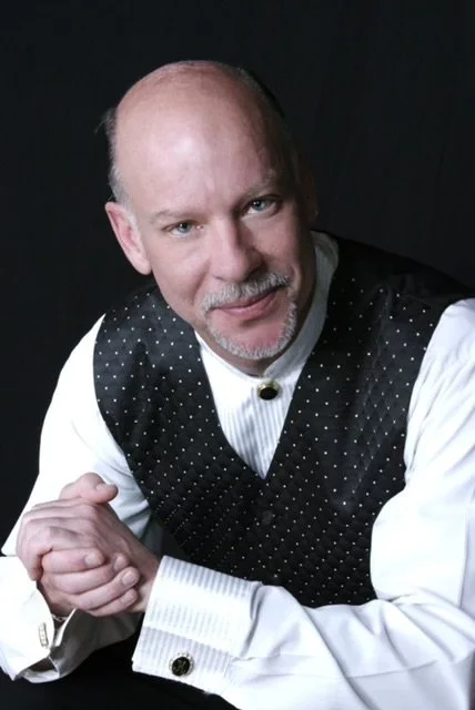 A middle-aged man with a bald head and a goatee, dressed in a white shirt with a black vest with white polka dots, smiling and looking at the camera against a black background.