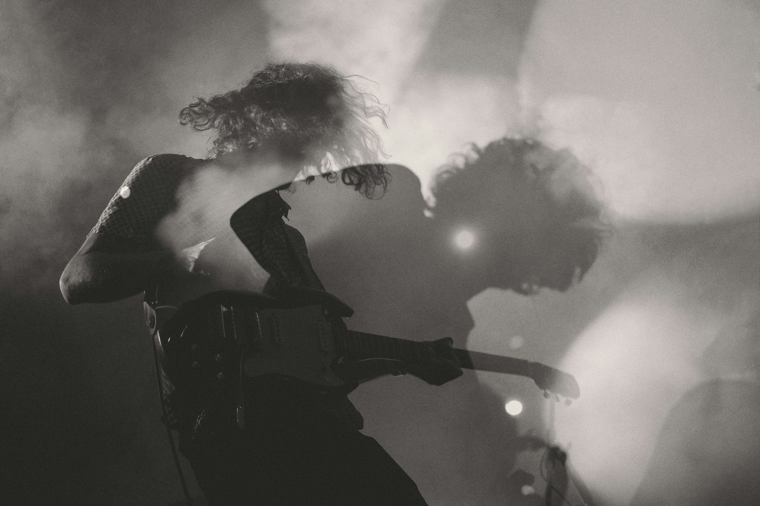 A silhouette of a person with curly hair, sitting in front of a smoky or foggy background, with sunlight or bright light in the distance.