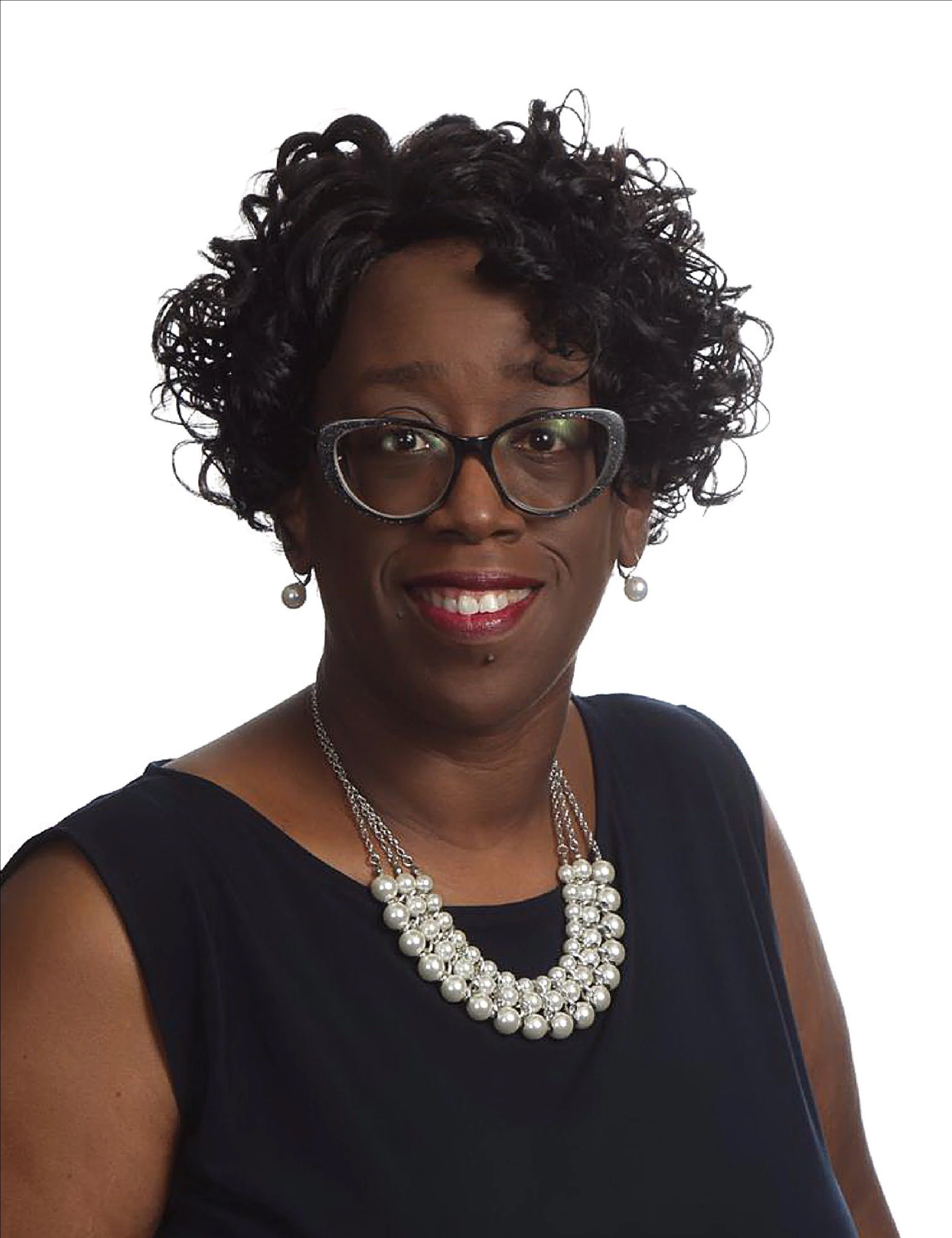 A woman with dark skin, short curly black hair, wearing glasses, a pearl necklace, pearl earrings, red lipstick, and a dark sleeveless top, smiling against a plain white background.