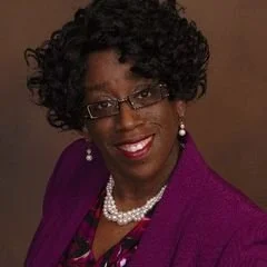 A woman with short, curly black hair, glasses, and earrings, smiling and wearing a purple jacket and pearl necklace.