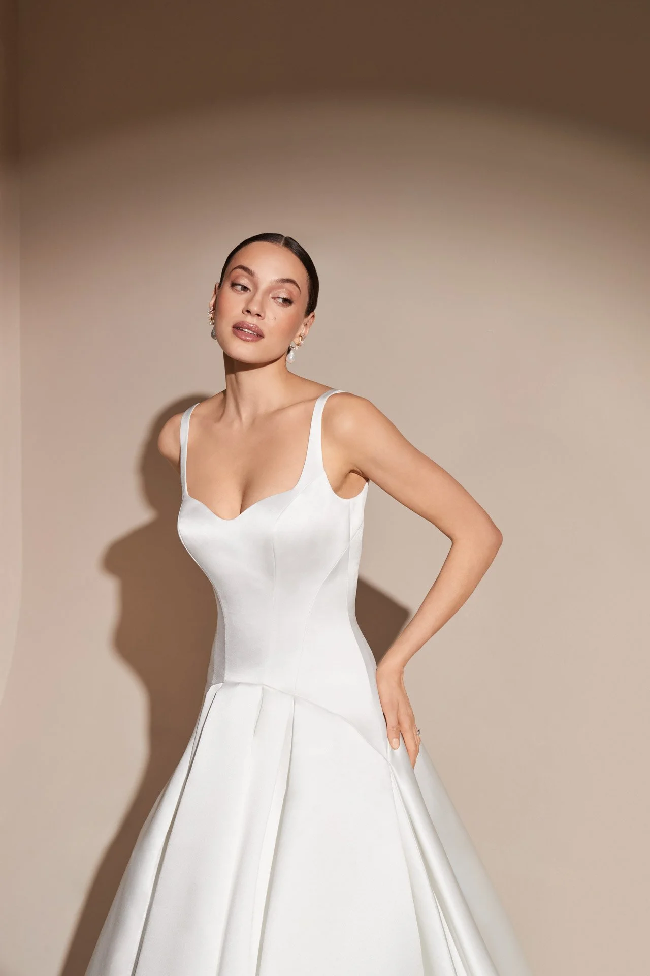 Close-up of a model wearing a structured Savannah Miller satin wedding dress featuring wide shoulder straps, a curved scoop neckline, and a dropped waist with soft pleating.