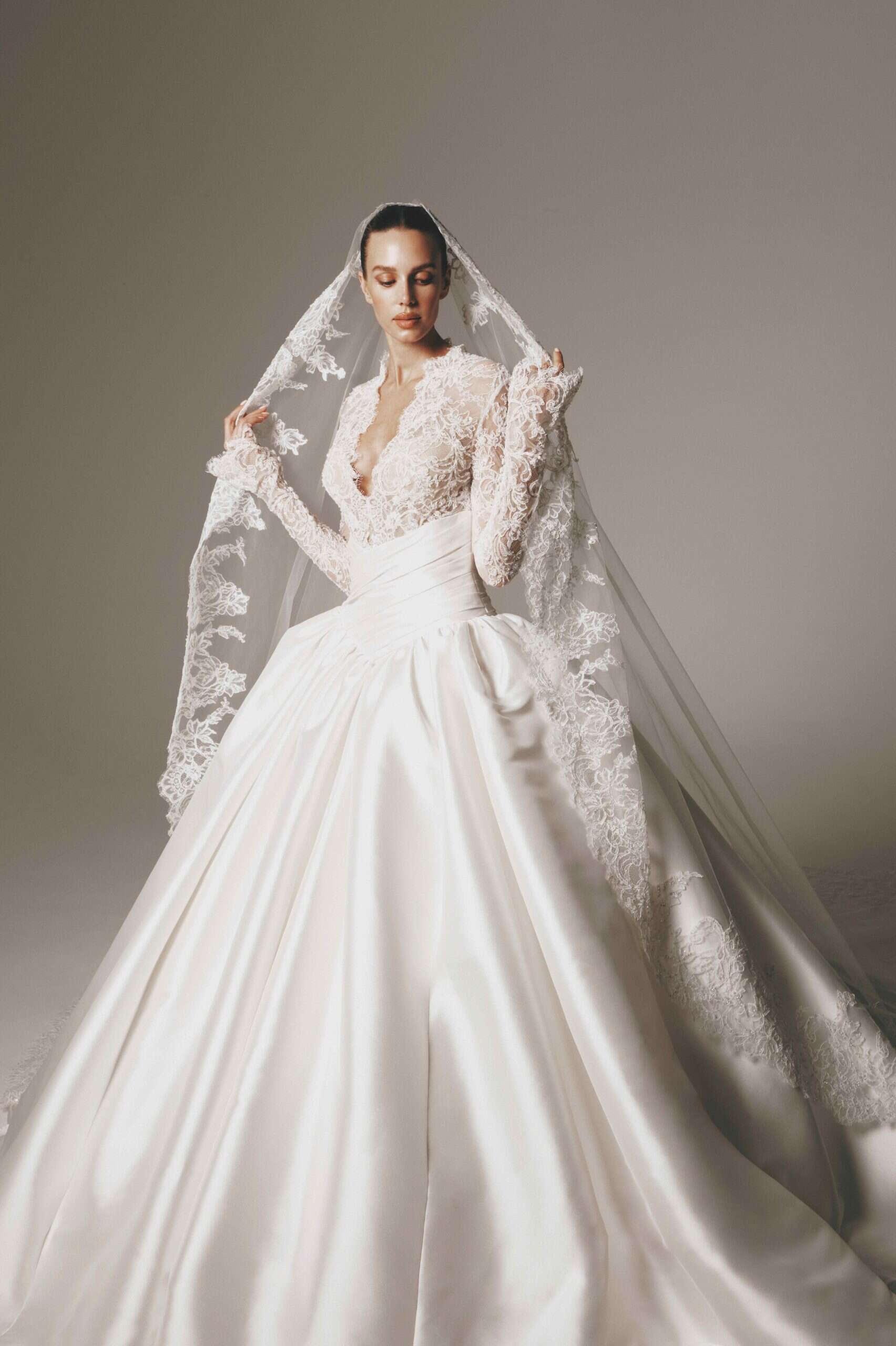 Woman in wedding dress with lace top and long pleated skirt, holding lace veil, against plain gray background.