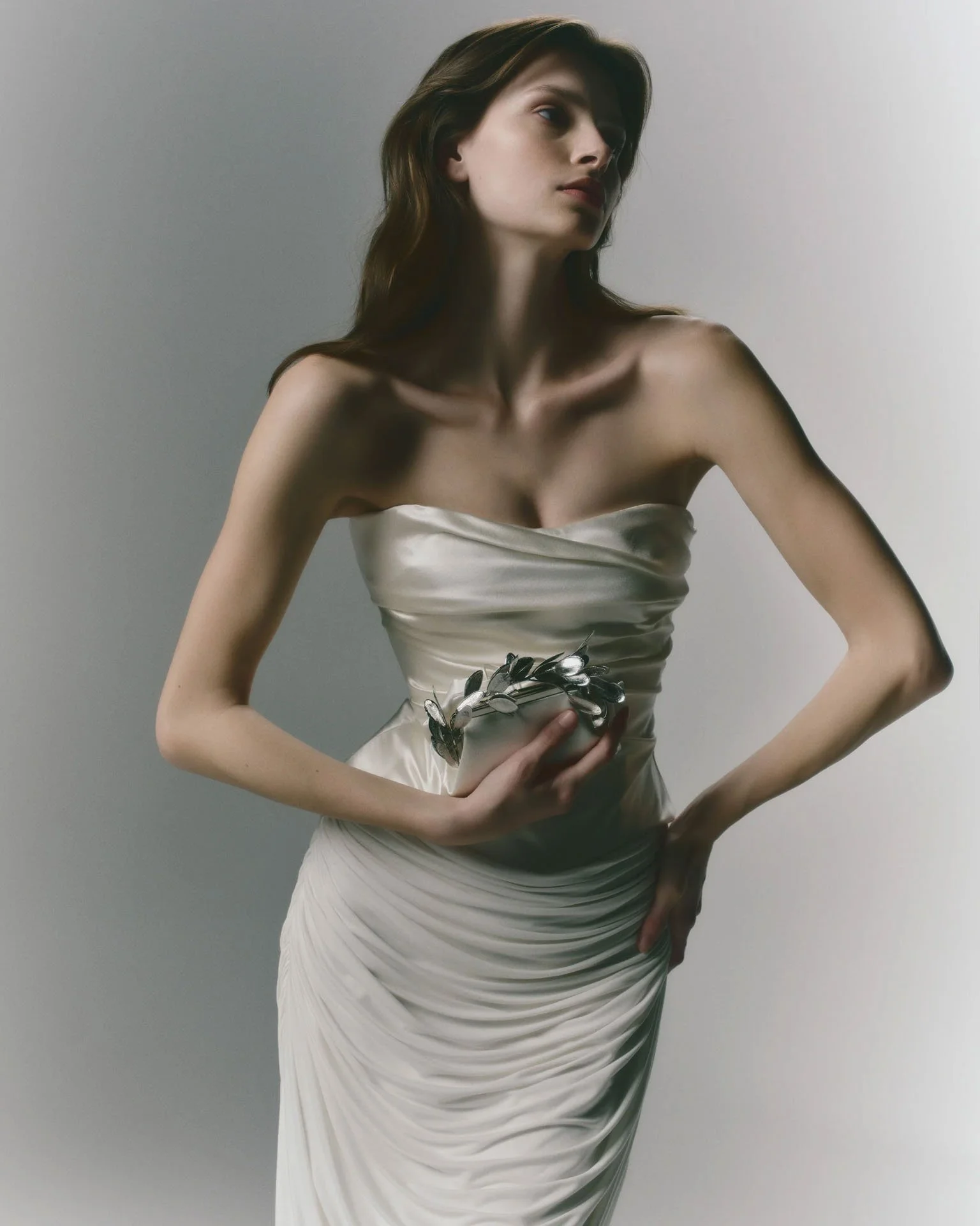 Model wearing a Danielle Frankel strapless, form-fitting wedding dress with intricate all-over ruching and draping, holding a sculpted silver floral bridal clutch.