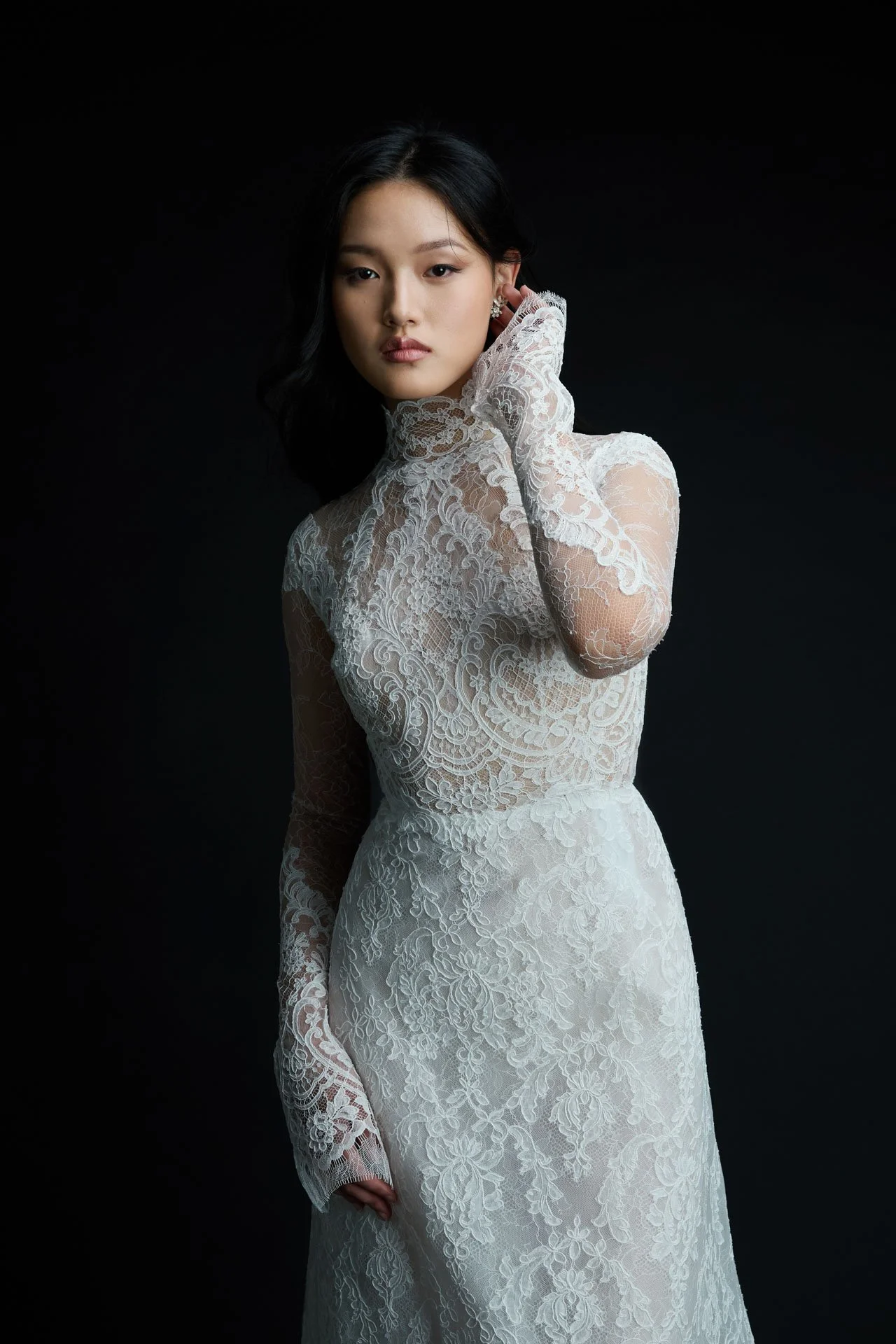 Close-up portrait of a model wearing a Jaclyn Whyte long-sleeve wedding dress highlighting a sheer illusion lace bodice, a high neckline, and delicate scalloped lace cuffs against a dark background.