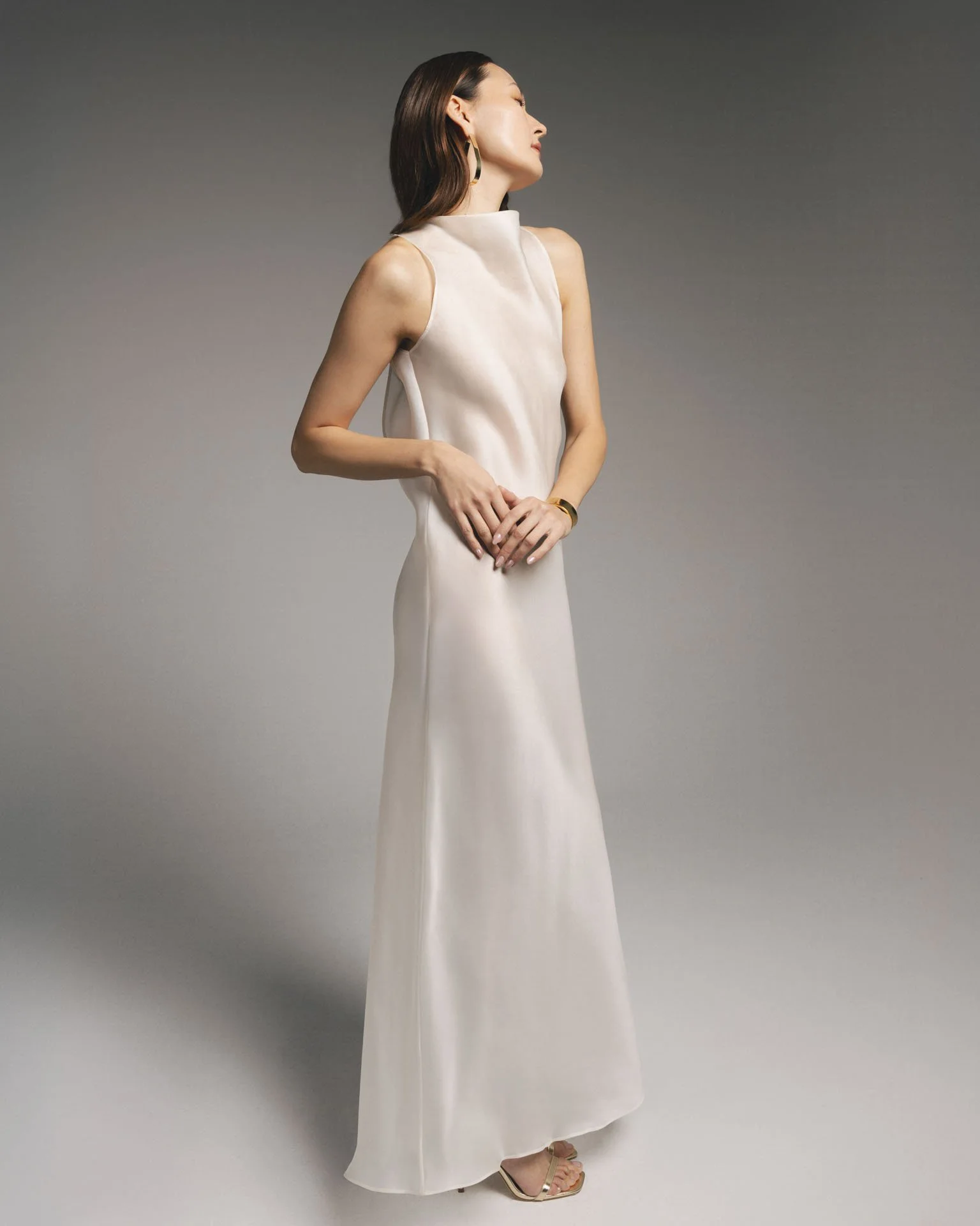 Model wearing a Karis minimalist off-white silk sleeveless maxi dress with a high neckline, paired with gold strappy heels and gold jewelry.