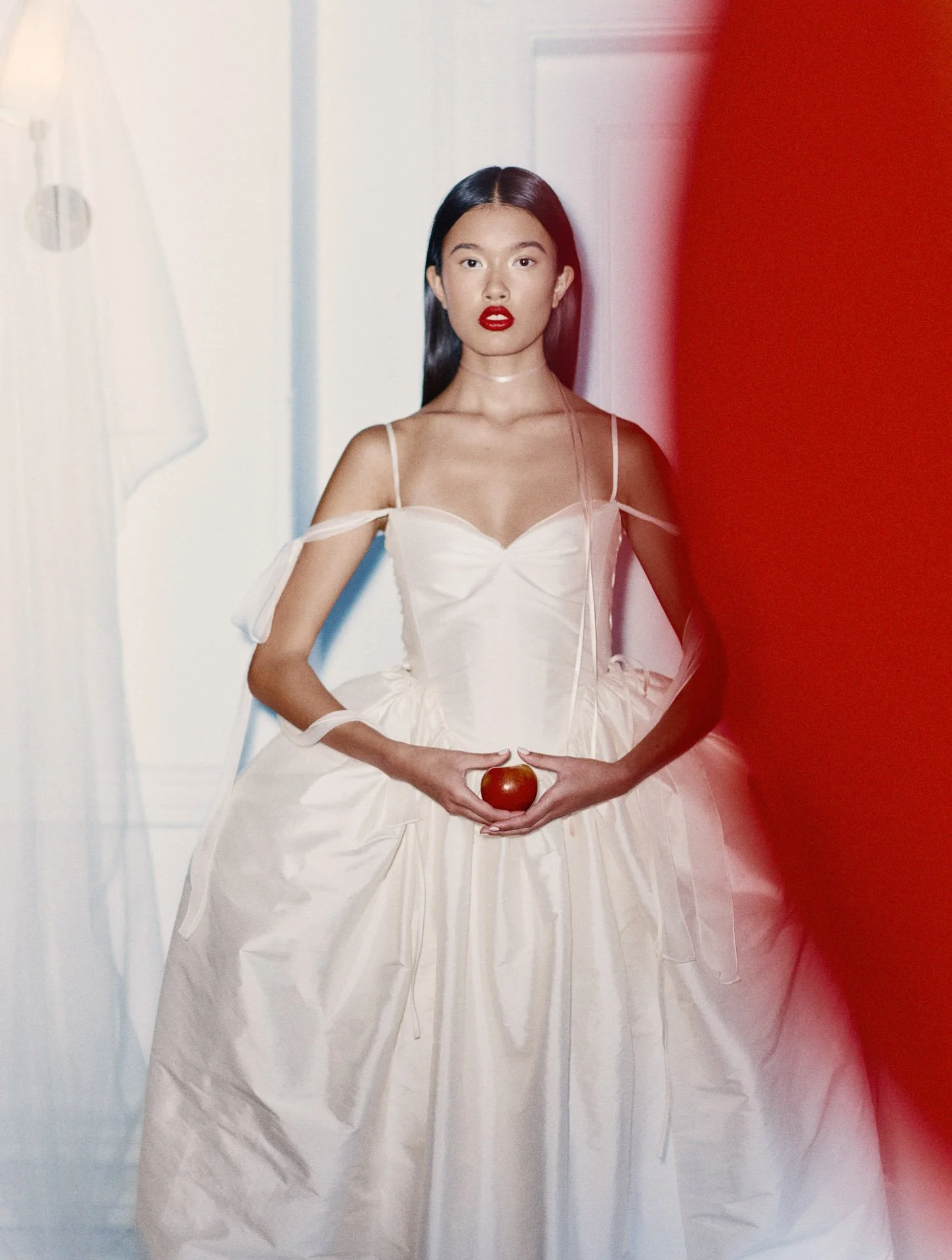 Model wearing a Former Girlfriend basque waist ballgown wedding dress featuring a sweetheart neckline, thin ribbon straps, and a highly voluminous taffeta skirt, holding a small red apple.