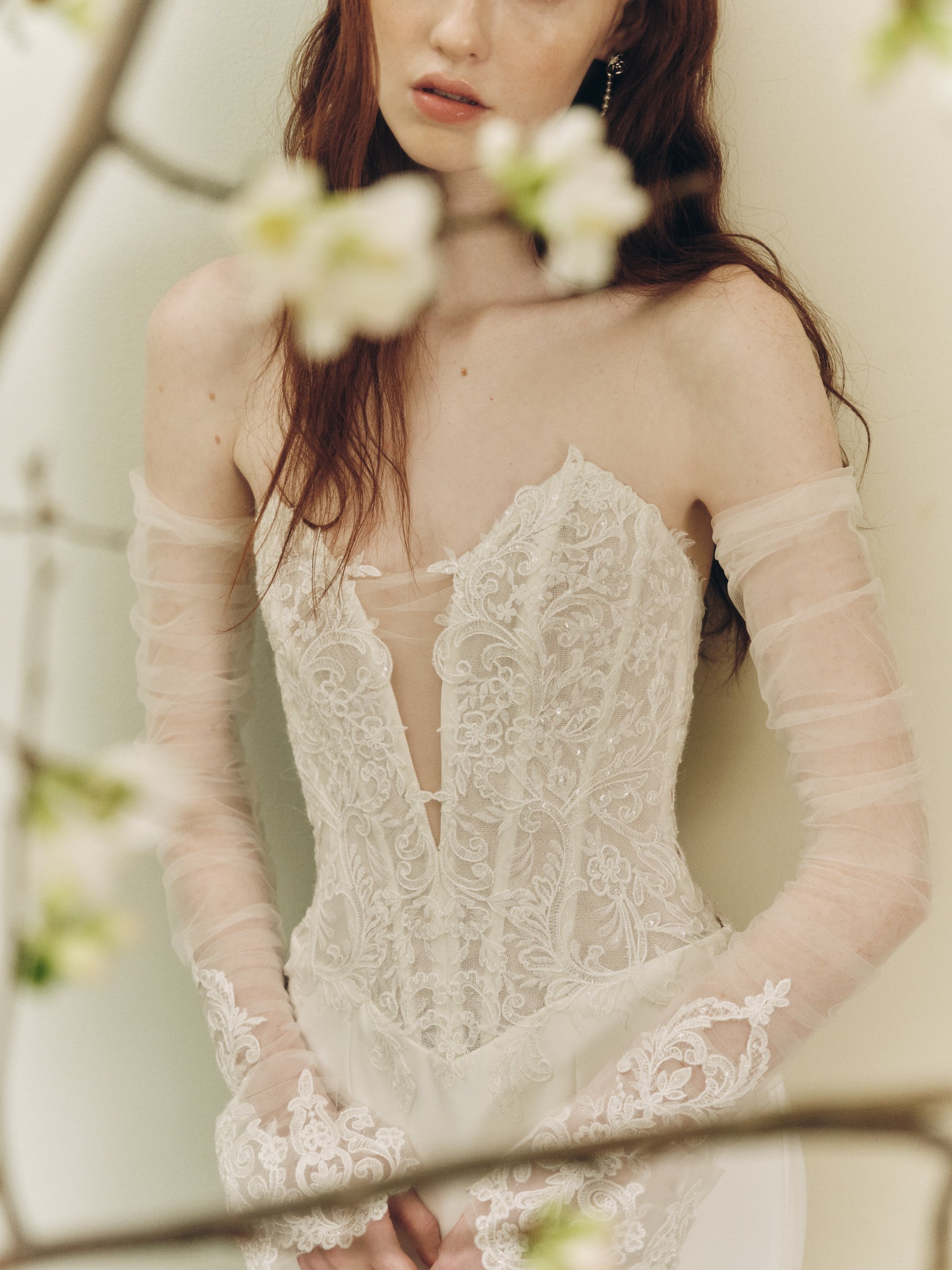 Close-up of a model wearing an Alyne strapless lace wedding dress featuring a deep illusion plunge neckline, intricate floral embroidery, and sheer detachable tulle sleeves with lace cuffs.