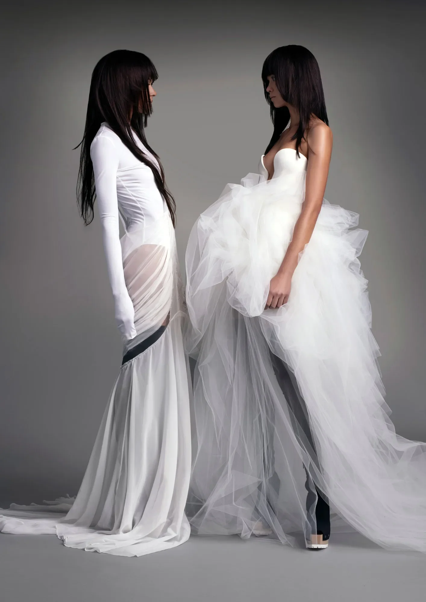 Two models showcasing modern, edgy Vera Wang wedding dresses; one in a sheer fitted gown over a long-sleeve white bodysuit, and the other in a strapless gown with a deep plunge and a highly voluminous tulle high-low skirt, both styled with black boot