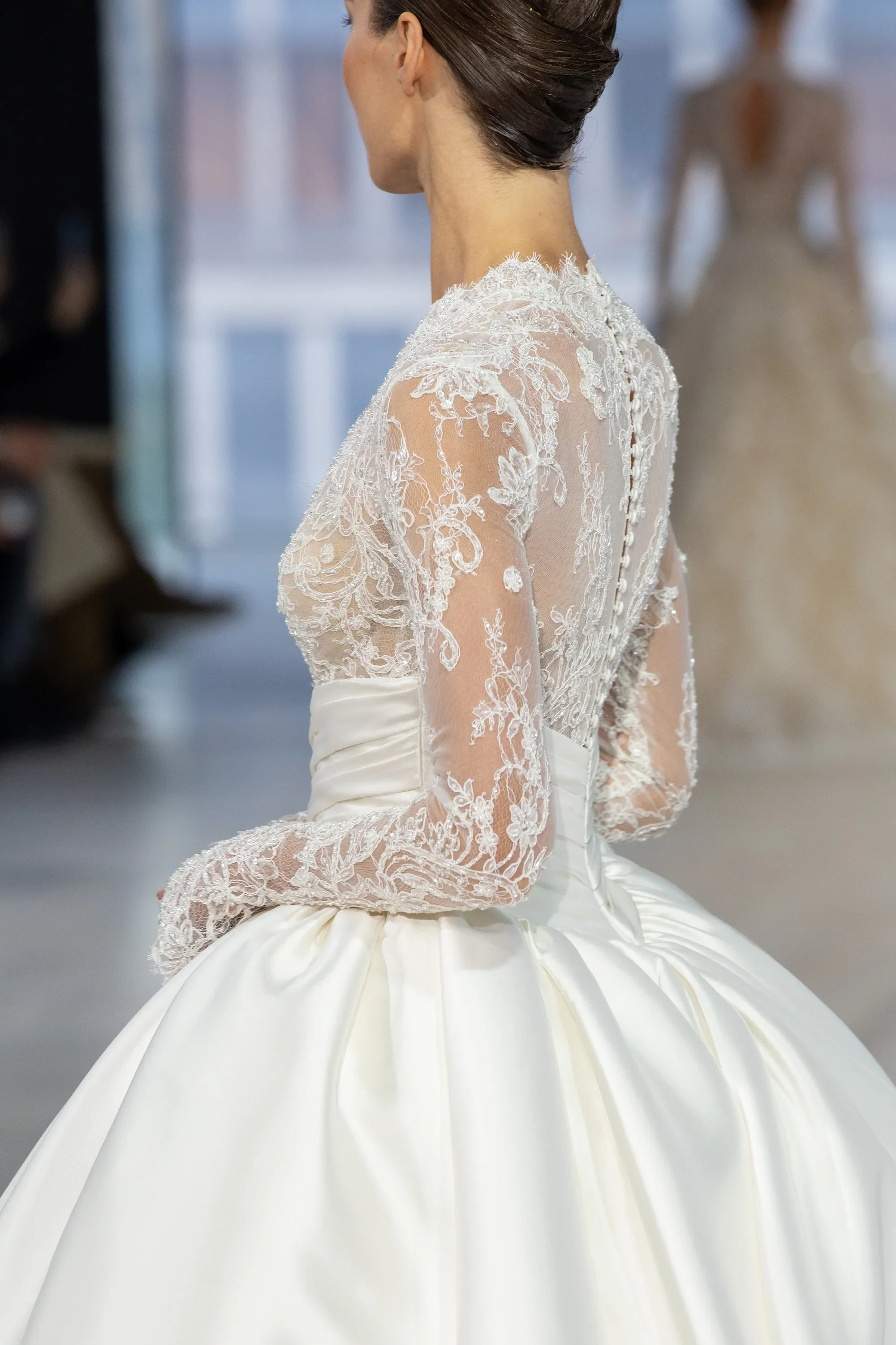 Side profile of a model wearing a Nicole and Felicia ballgown wedding dress showcasing a high-neck, long-sleeve sheer lace bodice with an elegant button-up back, paired with a voluminous, smoothly pleated satin skirt.