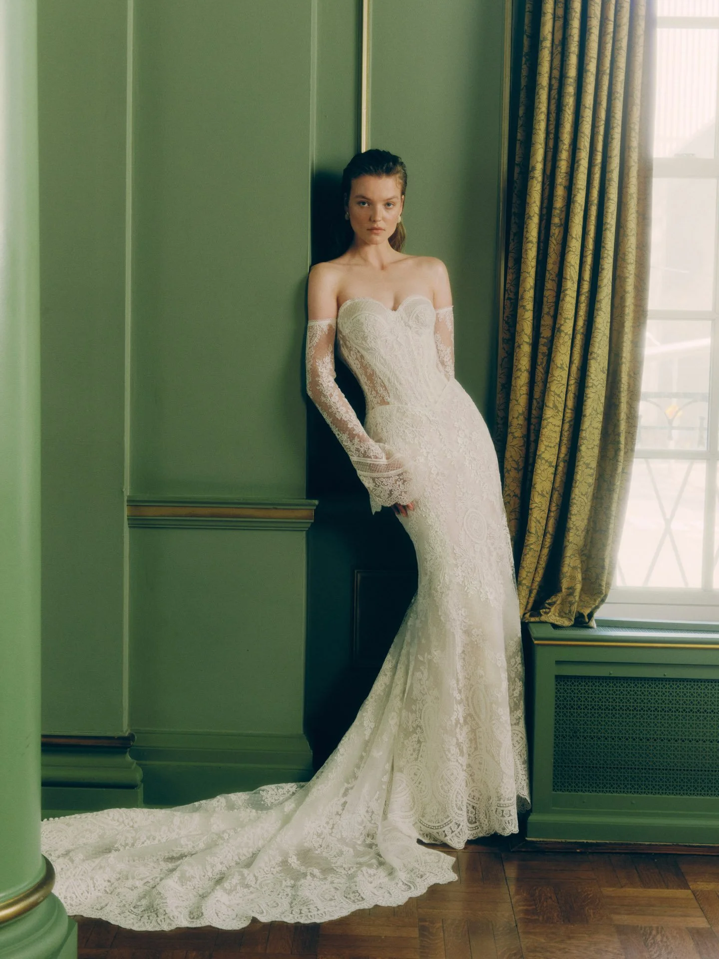 Model wearing a fitted Rivini strapless lace wedding dress featuring a sweetheart neckline, a sweeping scalloped lace train, and separate long flared lace sleeves, leaning against a dark green paneled wall