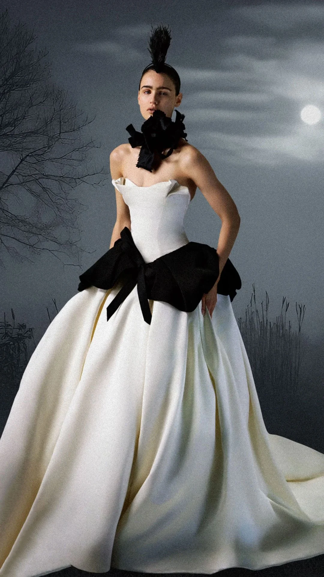 Model wearing a Vera Wang Haute strapless ballgown wedding dress featuring an architectural pointed bodice, a dramatic oversized black bow at the waist, and a bold black floral neckpiece, standing in a misty, moonlit field.