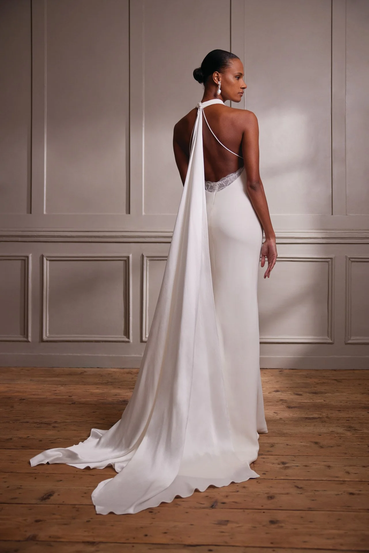 Back view of a model wearing a sleek Savannah Miller halter wedding dress, showcasing a plunging open back with scalloped lace detailing and a dramatic, flowing neck scarf that doubles as a train.
