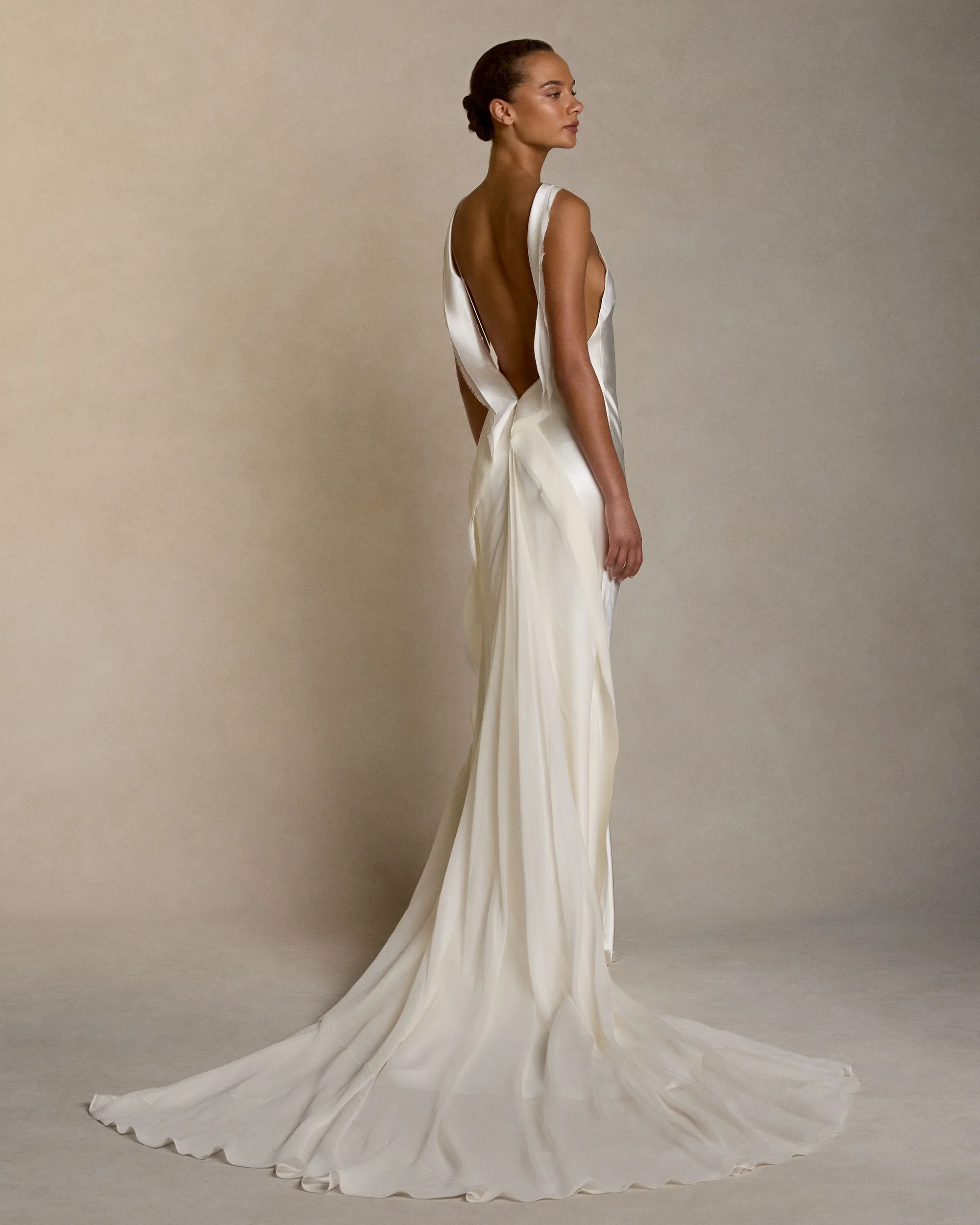 Back view of a model wearing a Danielle Frankel silk slip wedding dress, highlighting a plunging draped open back and an ethereal flowing sheer train.