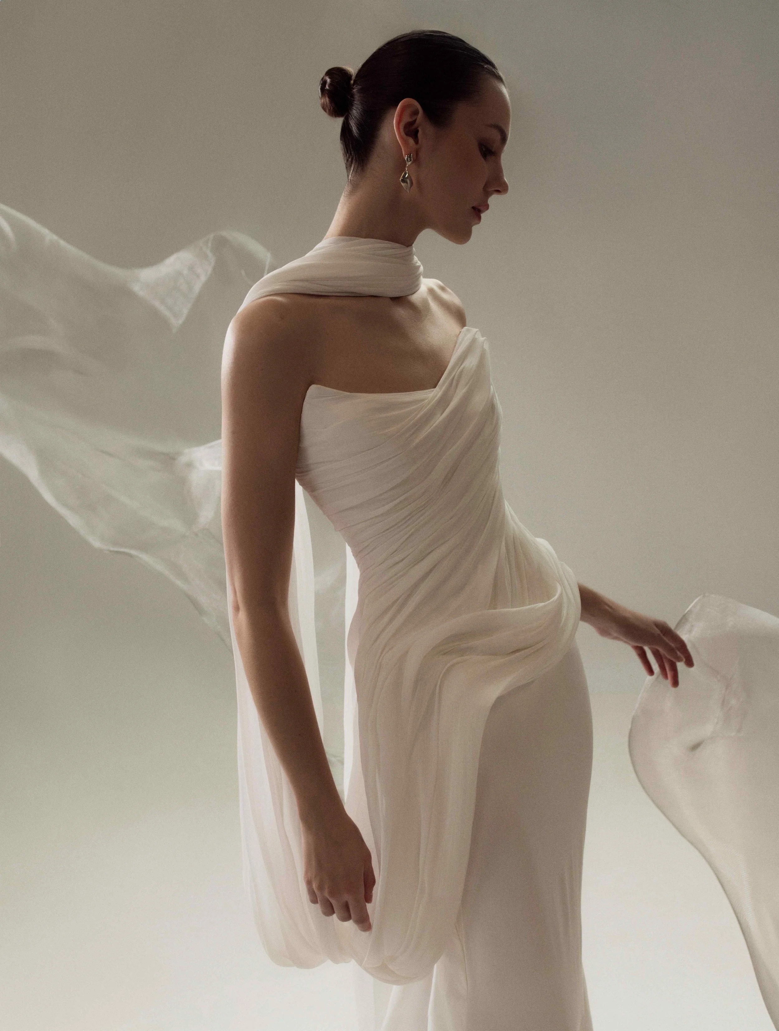 Model wearing a modern OUMA strapless wedding dress with elegant, sculptural asymmetrical draping across the bodice and a matching sheer neck scarf blowing in the wind.