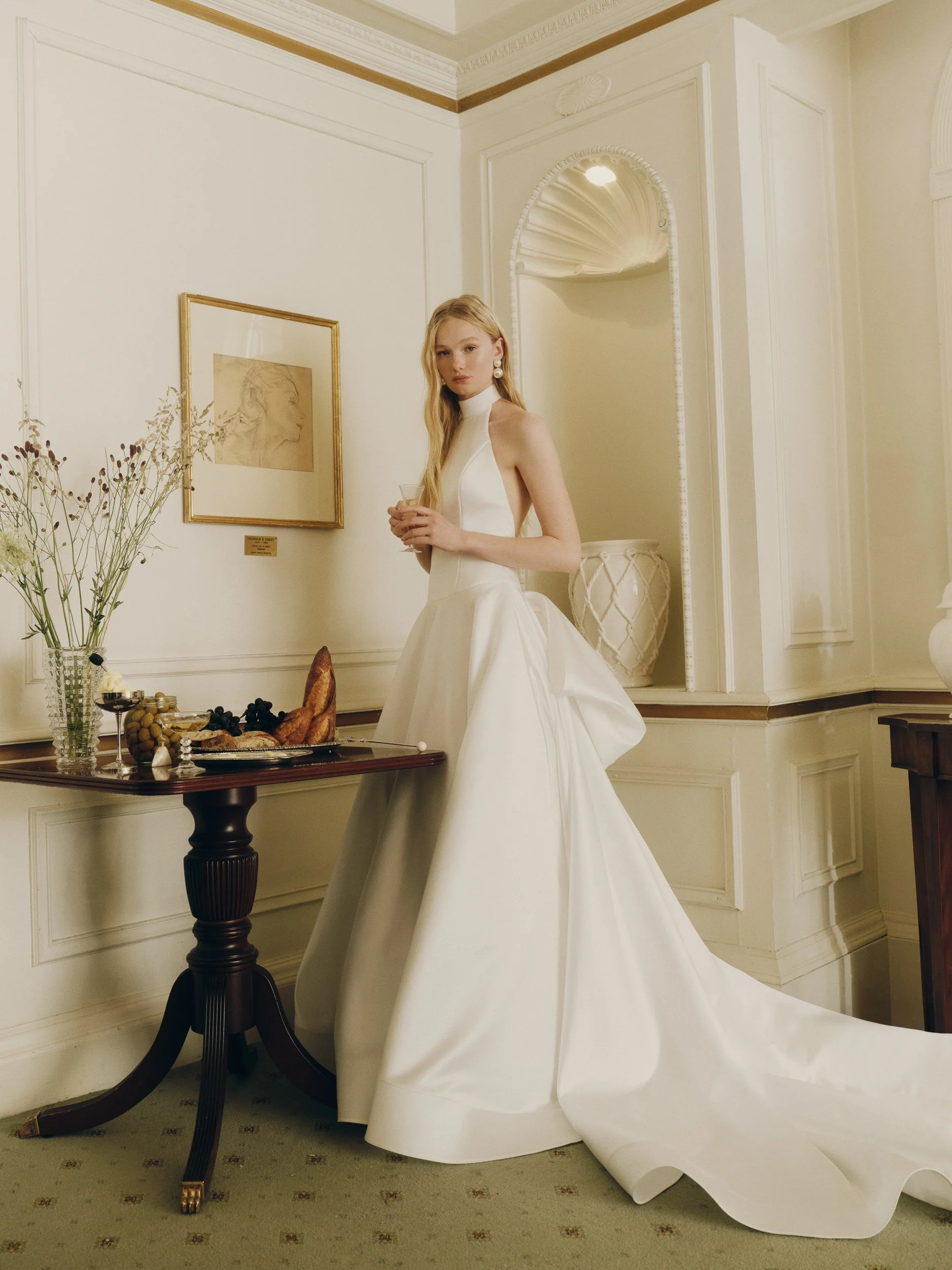 Model wearing the Alyne Pippa halter neck wedding dress, featuring a minimalist white bodice, an open back, and a dramatic sweeping train, standing in an elegant room.