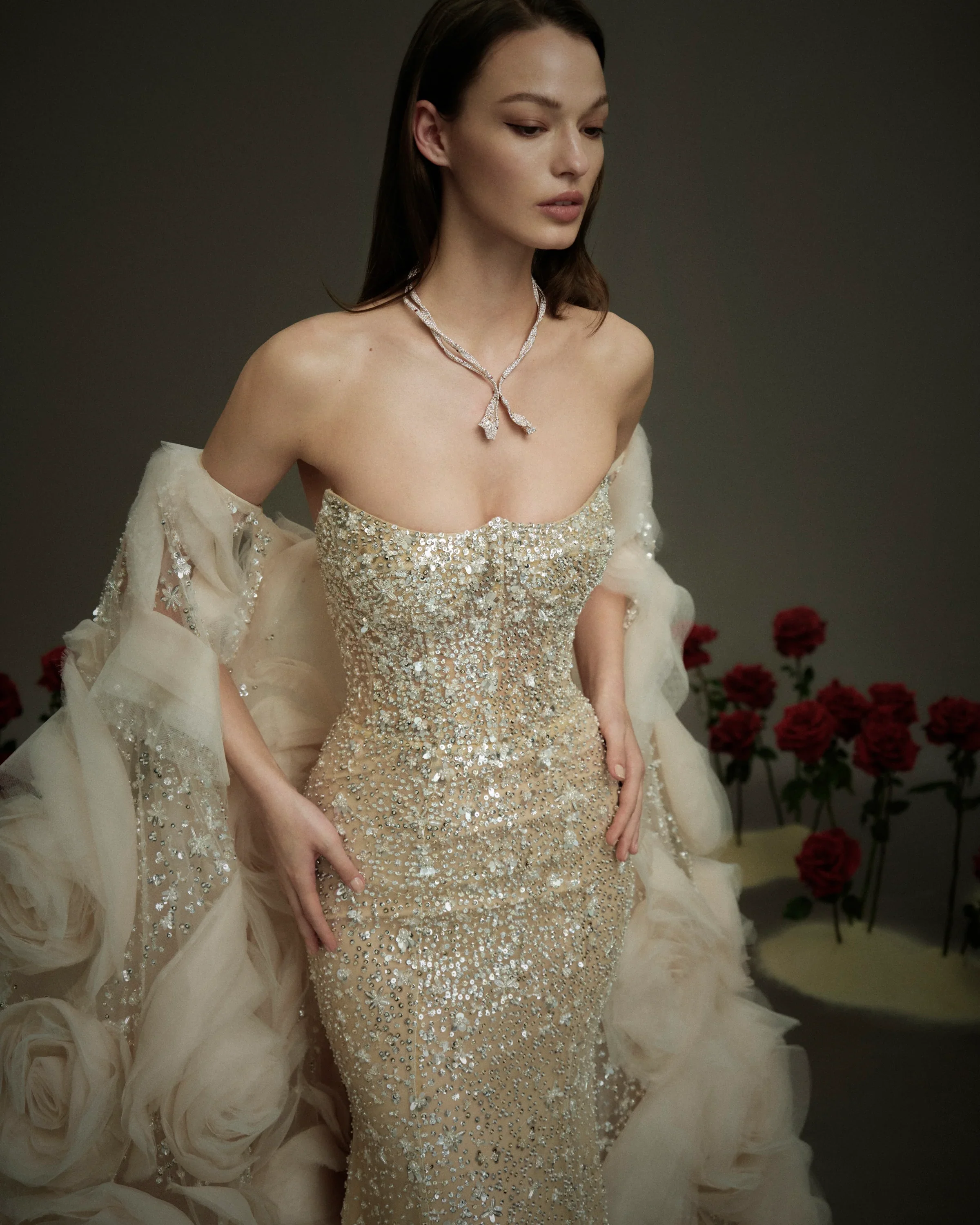 Model wearing a glamorous Nicole and Felicia strapless fitted wedding dress fully embellished with intricate silver sequins and crystal beadwork, paired with a dramatic ruffled tulle cape, standing near red roses.