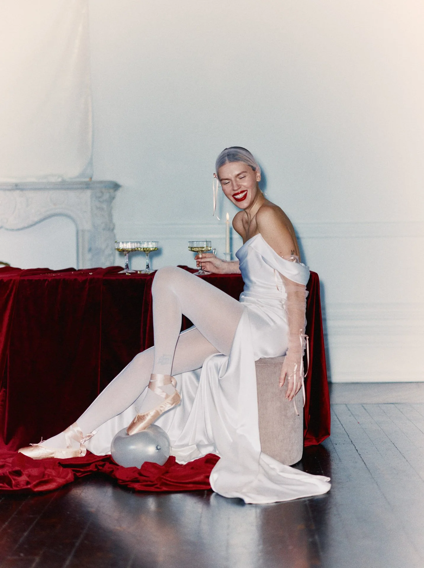 Model wearing a Former Girlfriend draped silk wedding dress featuring delicate off-the-shoulder sheer sleeves with ribbon ties, styled with ballet pointe shoes and white tights while holding a champagne glass.