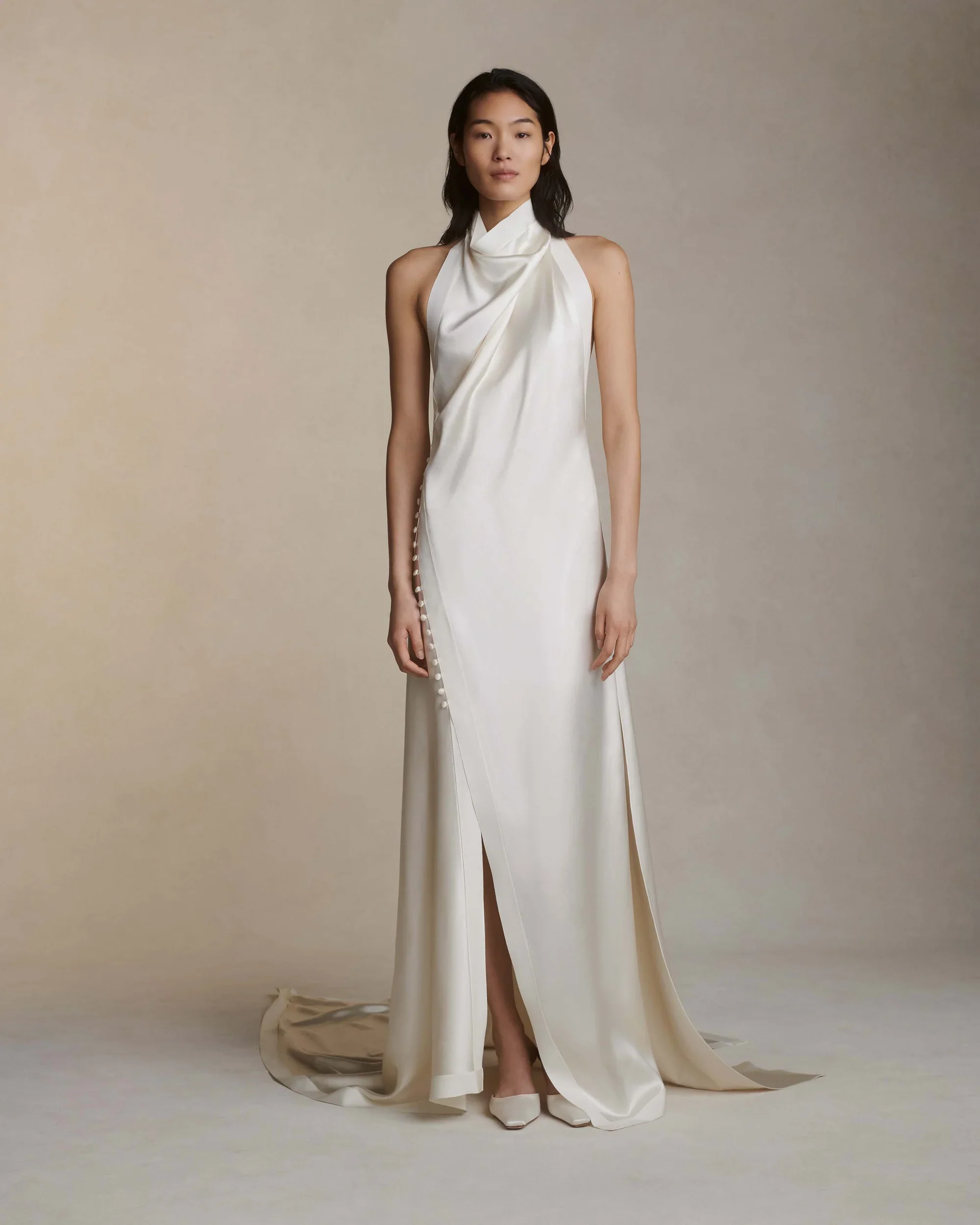 Model wearing a Danielle Frankel draped satin halter neck wedding dress featuring an asymmetrical wrap design, a high side slit detailed with delicate buttons, and a sweeping train.