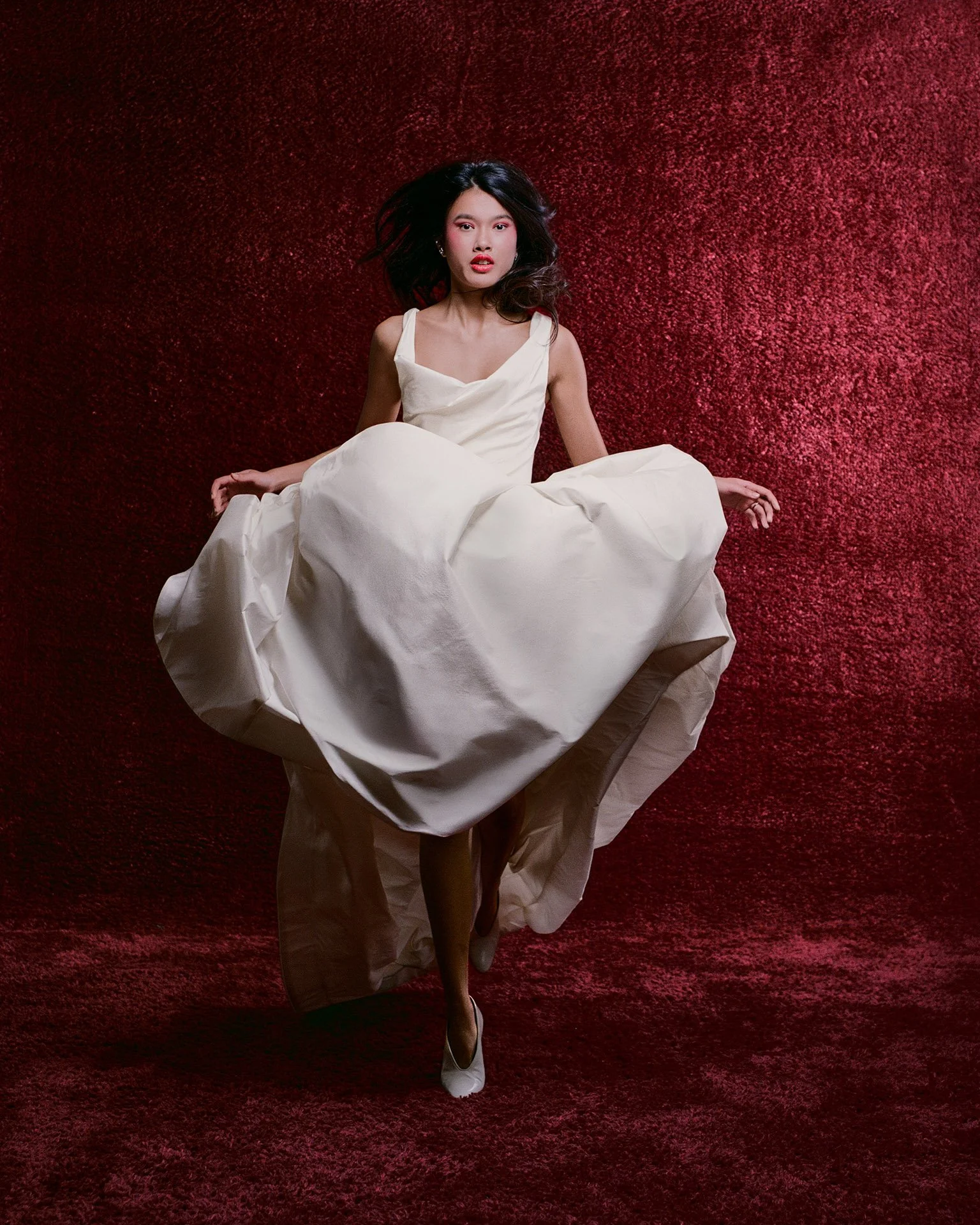 Model wearing a Former Girlfriend sleeveless wedding dress featuring a scoop neckline and a full voluminous skirt, playfully holding up the fabric while stepping forward against a textured dark red background.