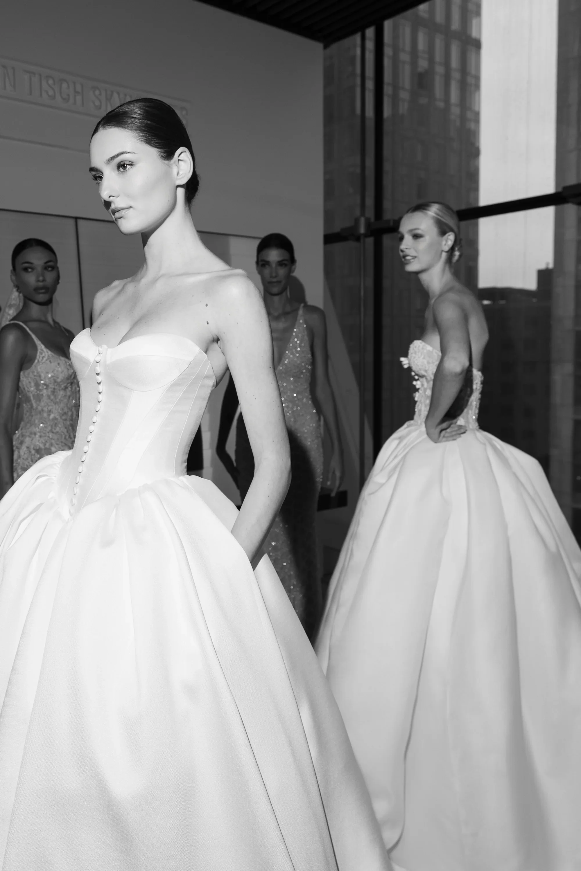 Black and white photograph of a model wearing a Nicole and Felicia strapless satin ballgown wedding dress featuring a sweetheart neckline, a button-front corset bodice with visible boning, and a full pleated skirt with pockets.