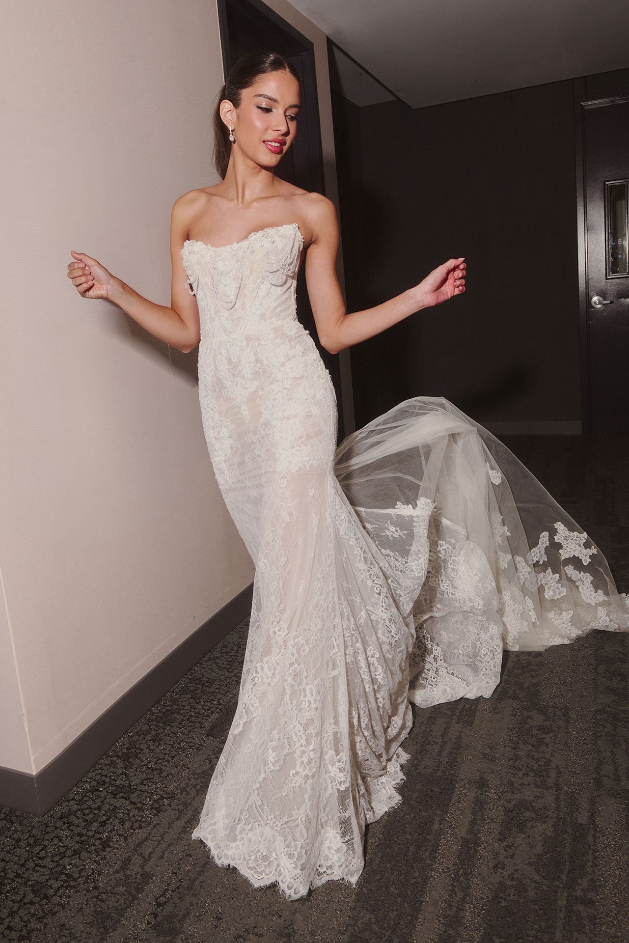 Model wearing a strapless Ines Di Santo fit-and-flare lace wedding dress featuring a unique draped scalloped neckline and a dramatic, flowing sheer lace train.