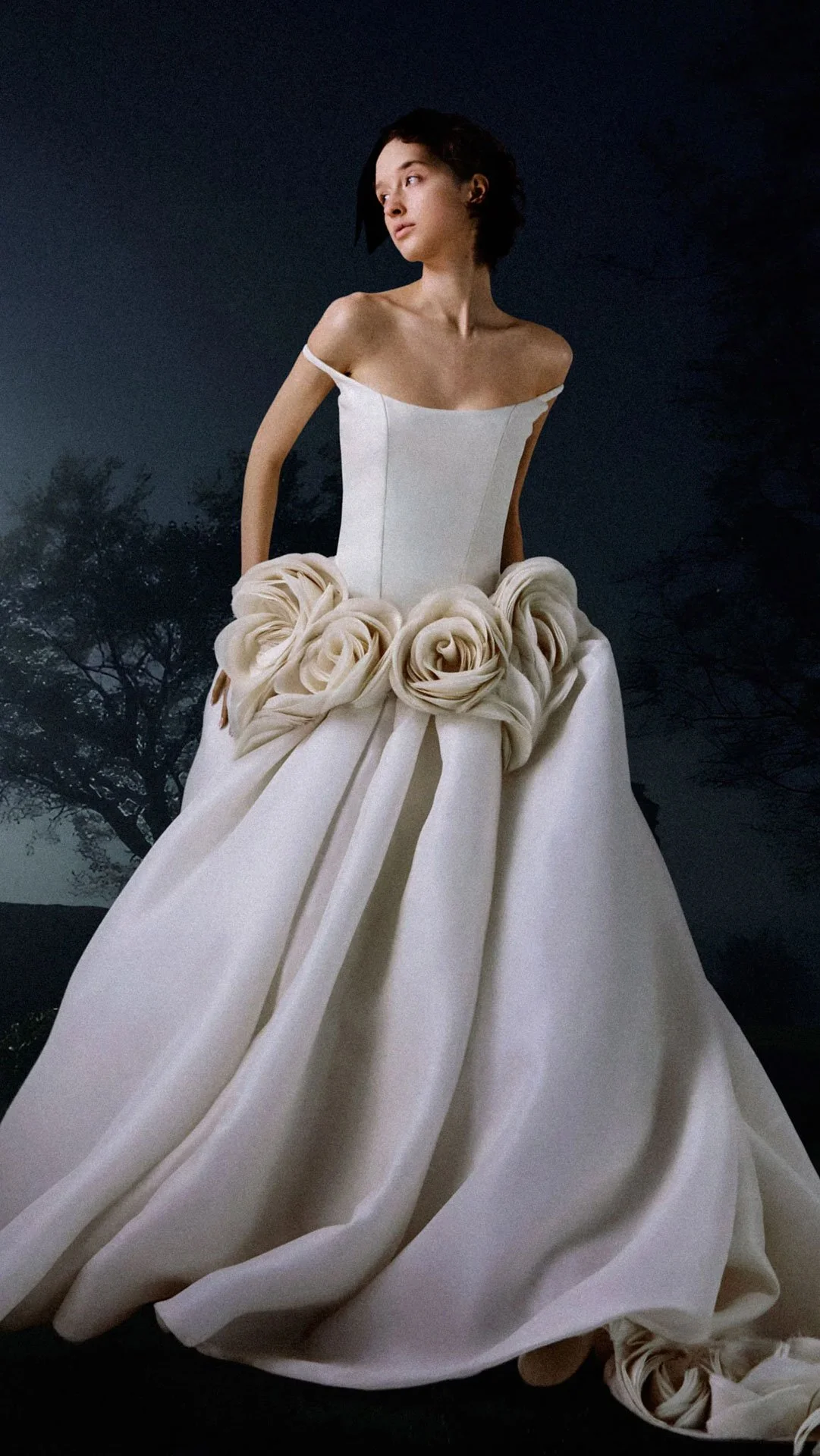 Model wearing a Vera Wang Haute off-the-shoulder wedding dress featuring a smooth, structured minimalist bodice and oversized sculpted fabric rosettes at the drop waist, flowing into a voluminous skirt.