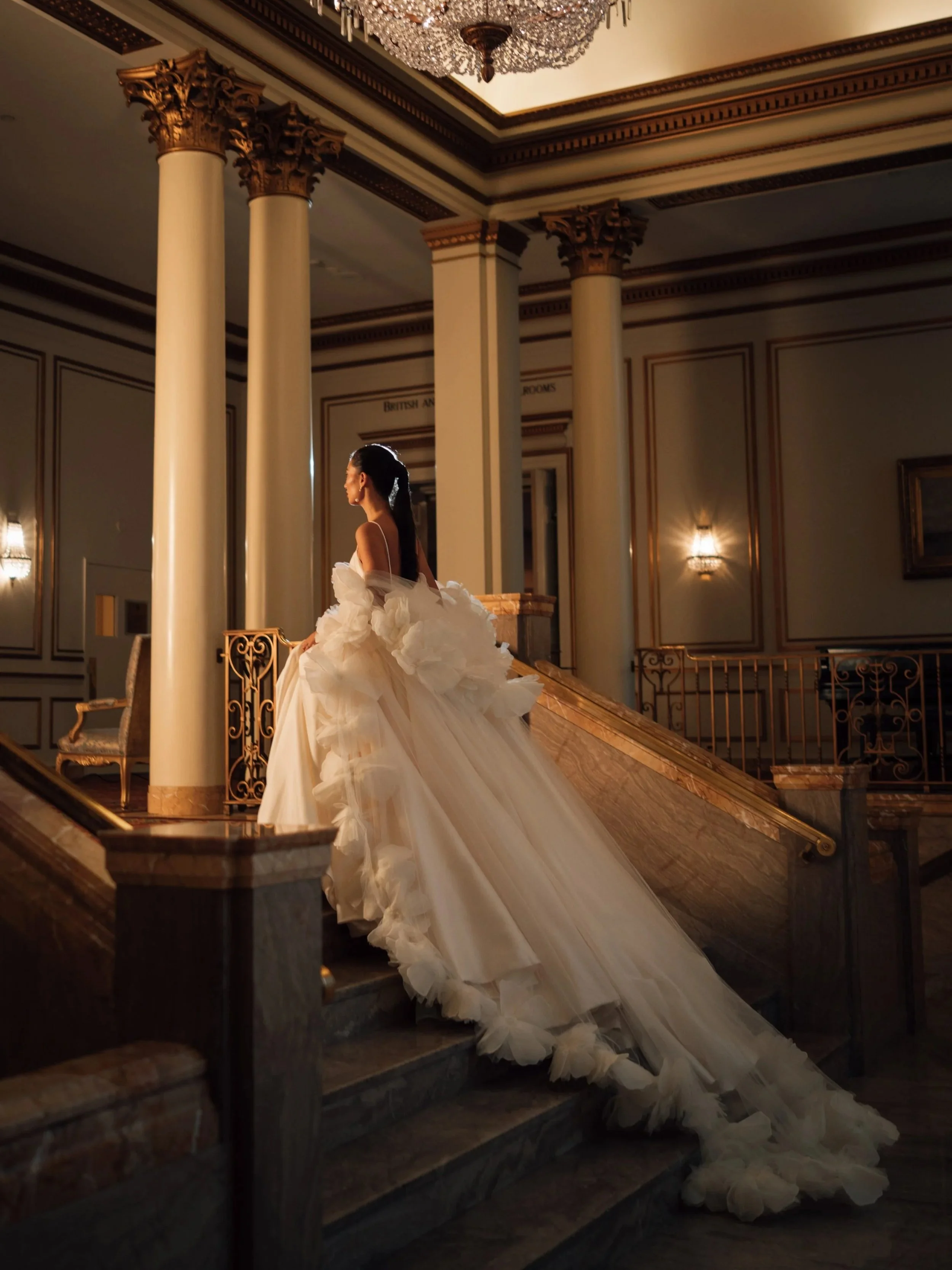 A woman in an elegant, voluminous white wedding gown with a long train stands on a grand staircase inside a luxurious building. The interior features tall columns, ornate gold accents, a large chandelier, and warm lighting.