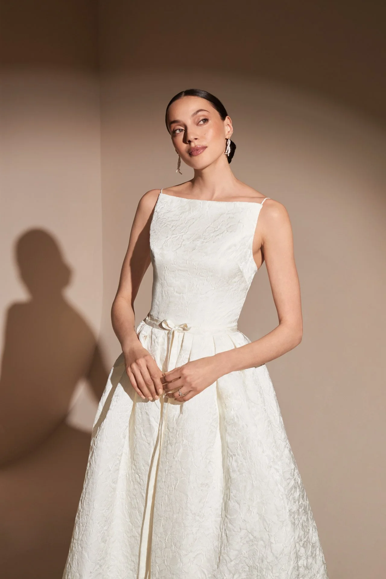 Close-up of a model wearing a Savannah Miller A-line wedding dress crafted from textured jacquard fabric, featuring a sleek straight neckline, thin straps, and a delicate ribbon tied at the waist.