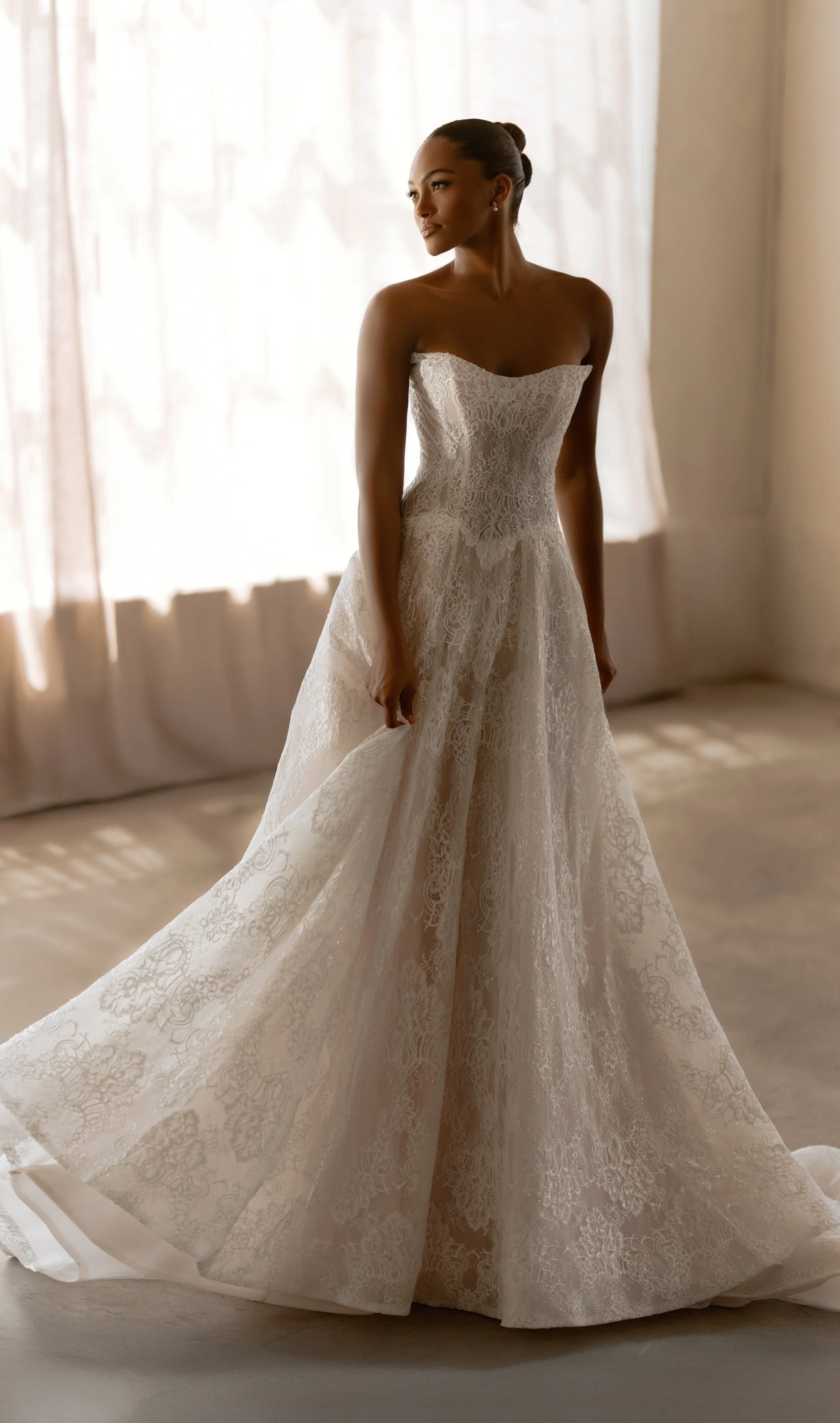 Model wearing a Martina Liana strapless A-line wedding dress made of all-over delicate lace, standing indoors by a softly lit window.