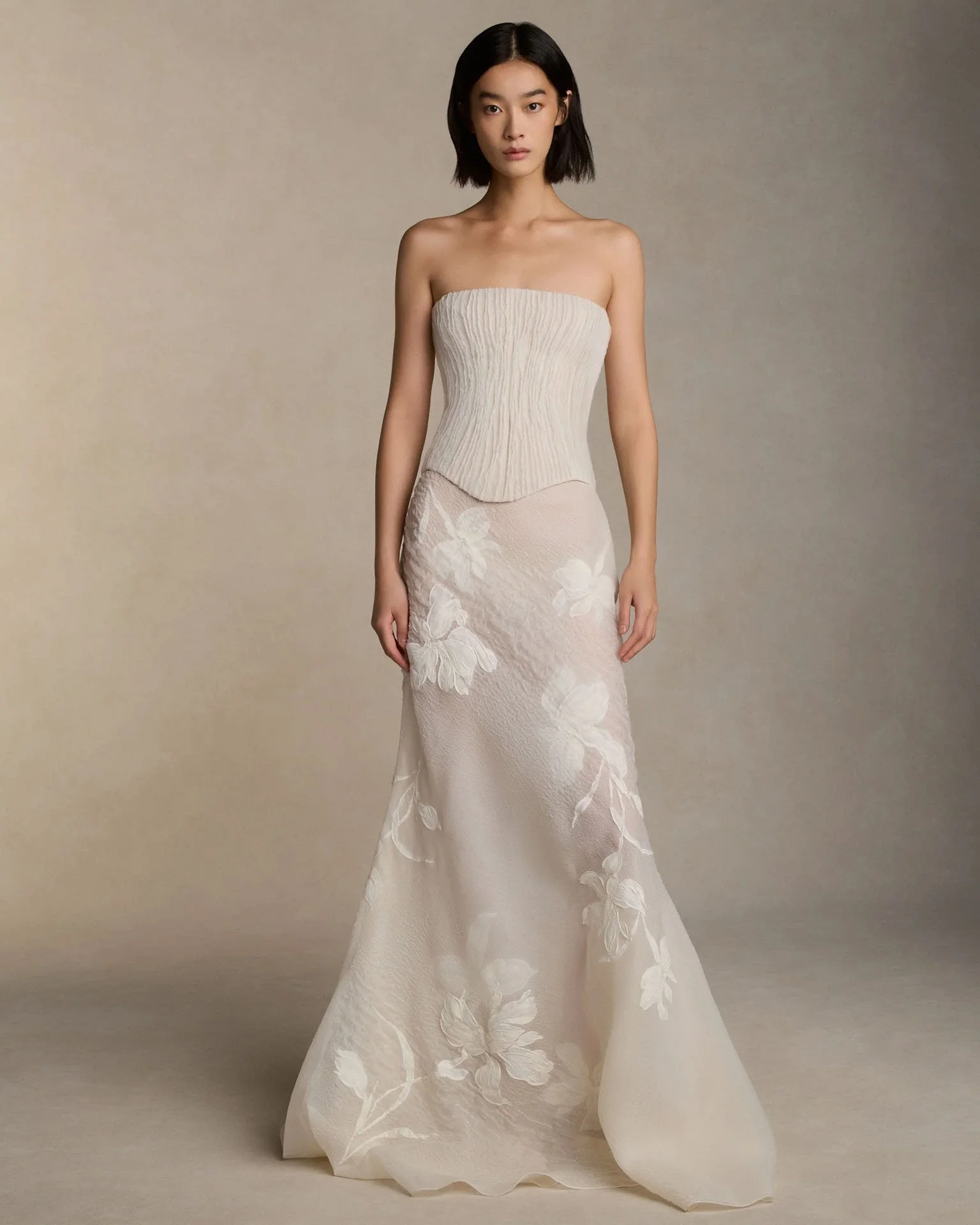 Model wearing a Danielle Frankel strapless wedding dress featuring a delicately pleated corset bodice and a sheer skirt adorned with oversized floral lace appliqués.