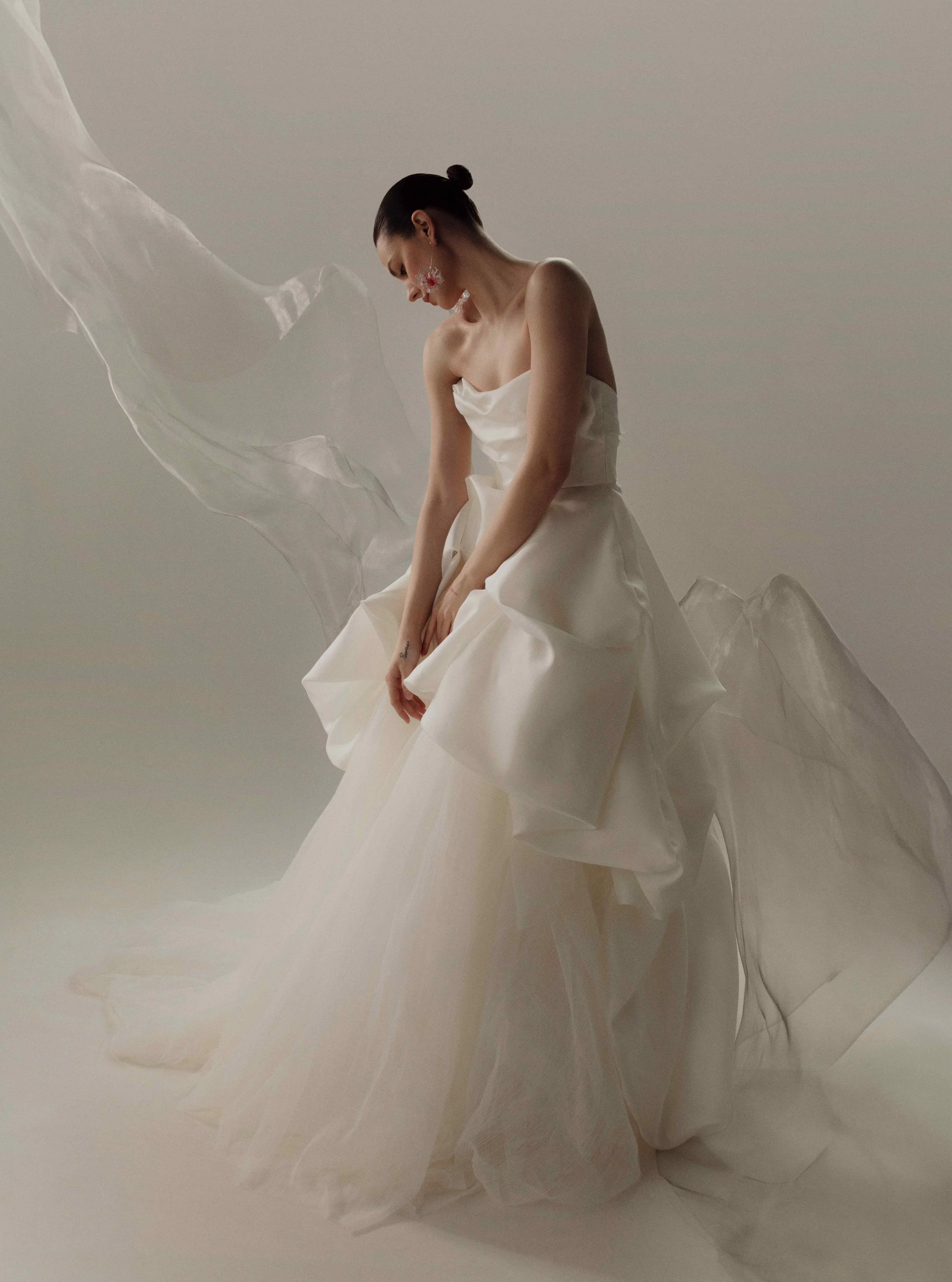 Model wearing an OUMA strapless wedding dress featuring architectural, dramatically draped tiers, standing with a sheer piece of lightweight fabric floating dynamically behind her.