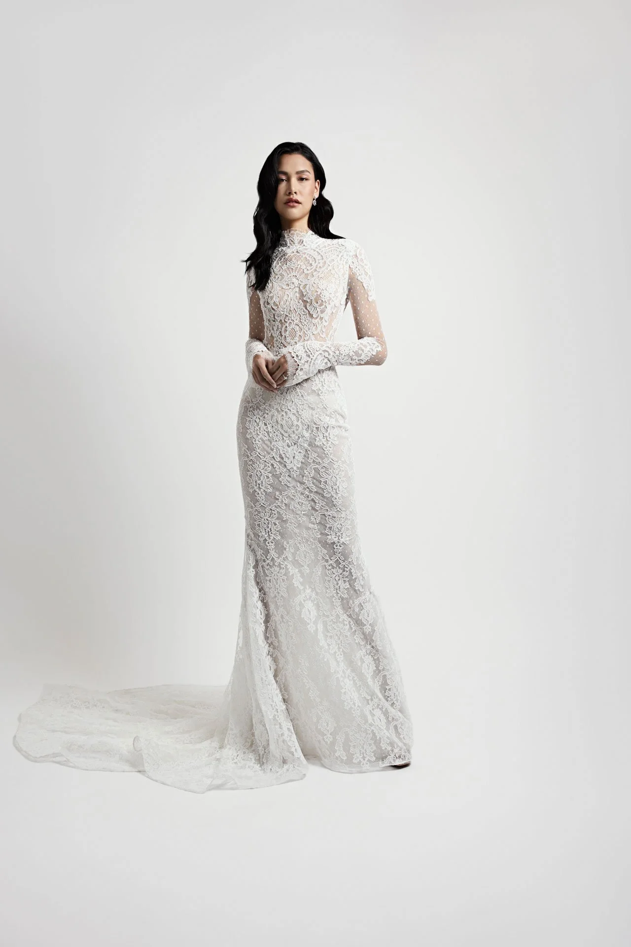Model wearing a Jaclyn Whyte fitted long-sleeve lace wedding dress featuring a sheer high mock neckline, intricate all-over floral lace, and a delicate sweeping train, standing against a white studio backdrop