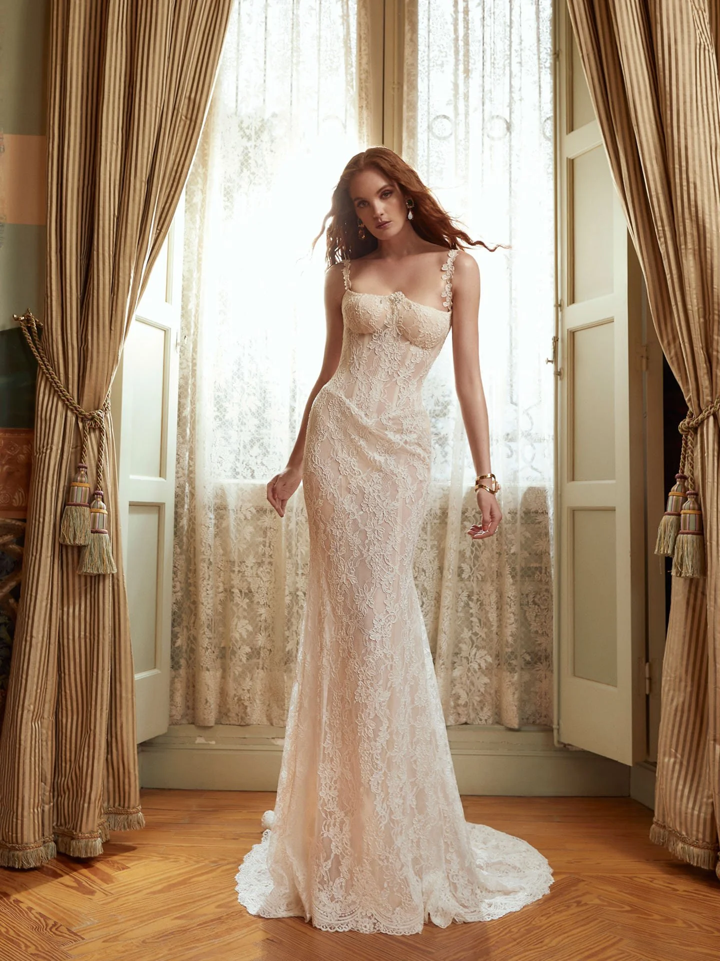 Model wearing a Galia Lahav Couture fit-and-flare lace wedding dress featuring a structured sheer corset bodice, delicate floral straps, and intricate all-over lace, standing in front of elegant window drapery.