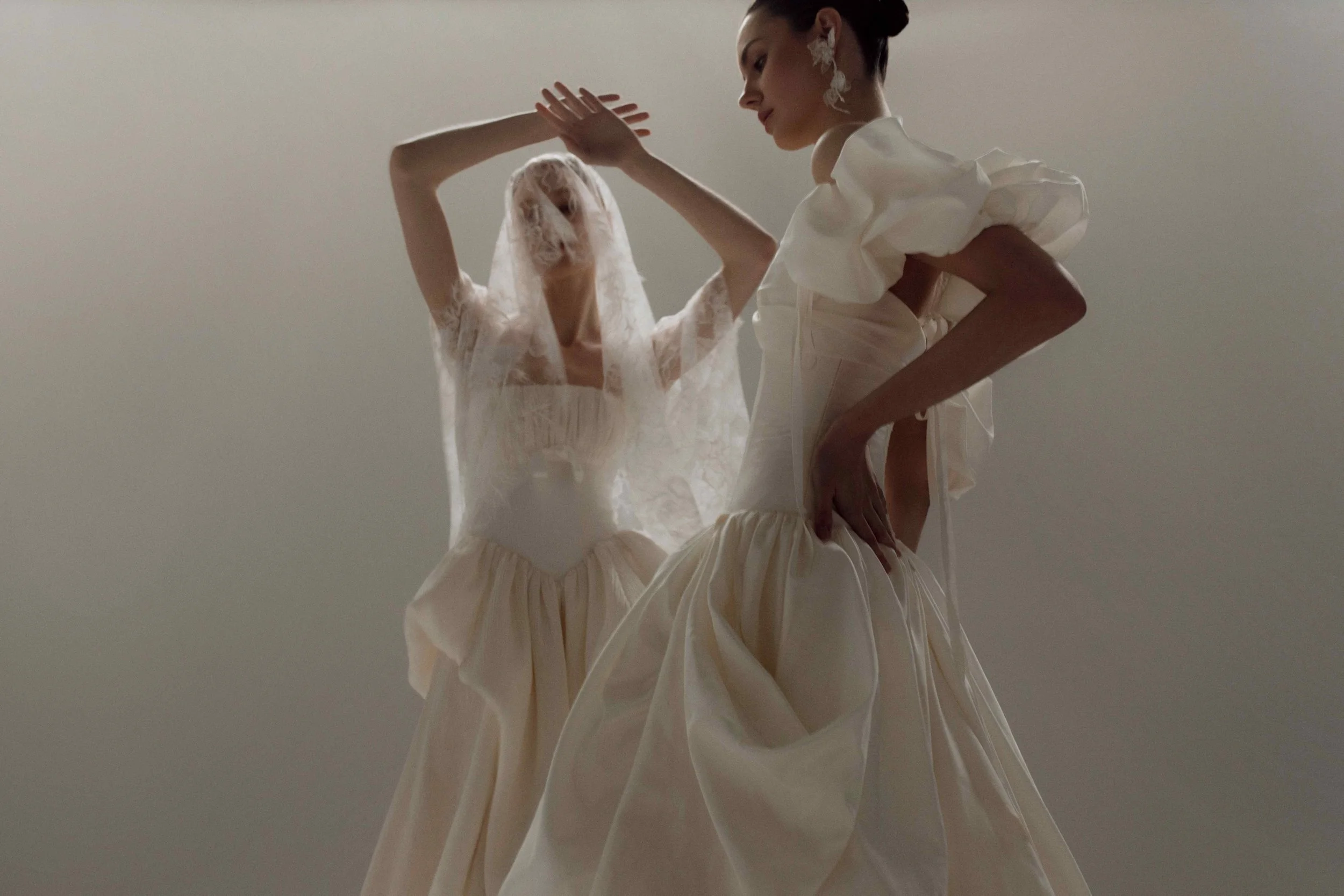 Two models showcasing OUMA romantic wedding dresses; one featuring voluminous puff sleeves and a structured drop waist, and the other wearing a delicate lace veil over a corseted gown.