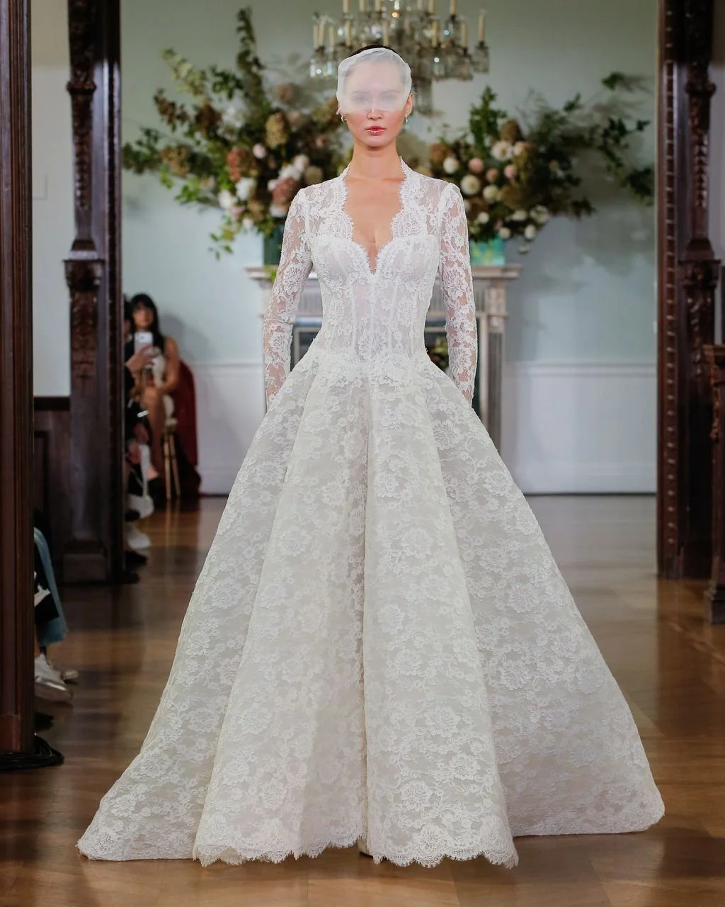 Model walking a runway in a Monique Lhuillier long-sleeve lace ballgown wedding dress featuring a plunging V-neckline, a structured corset bodice, and a full skirt, styled with a modern birdcage veil.
