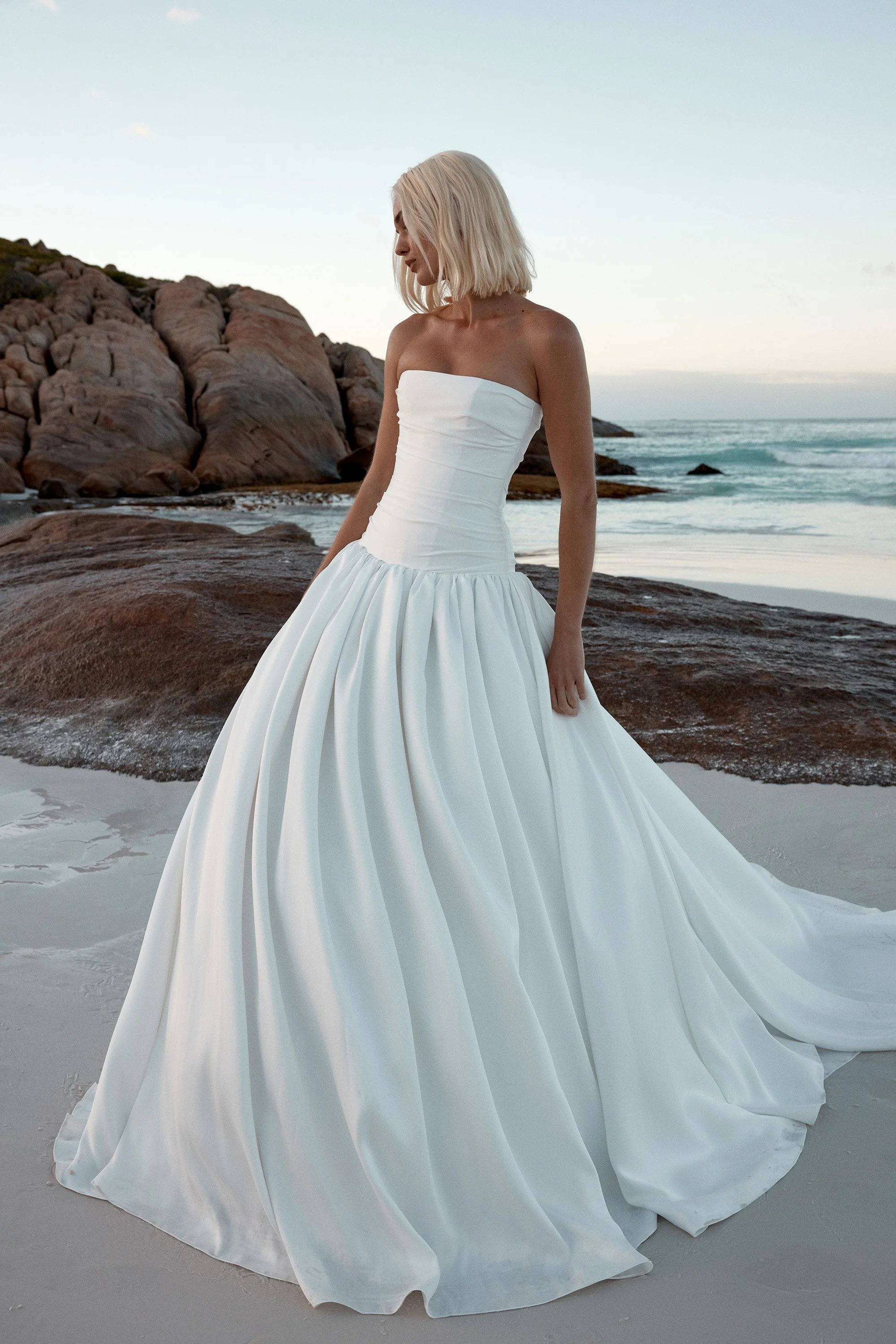 Model wearing a KYHA strapless wedding dress featuring a fitted, horizontally pleated drop-waist bodice that flows into a full gathered skirt, standing on a sandy beach at twilight.