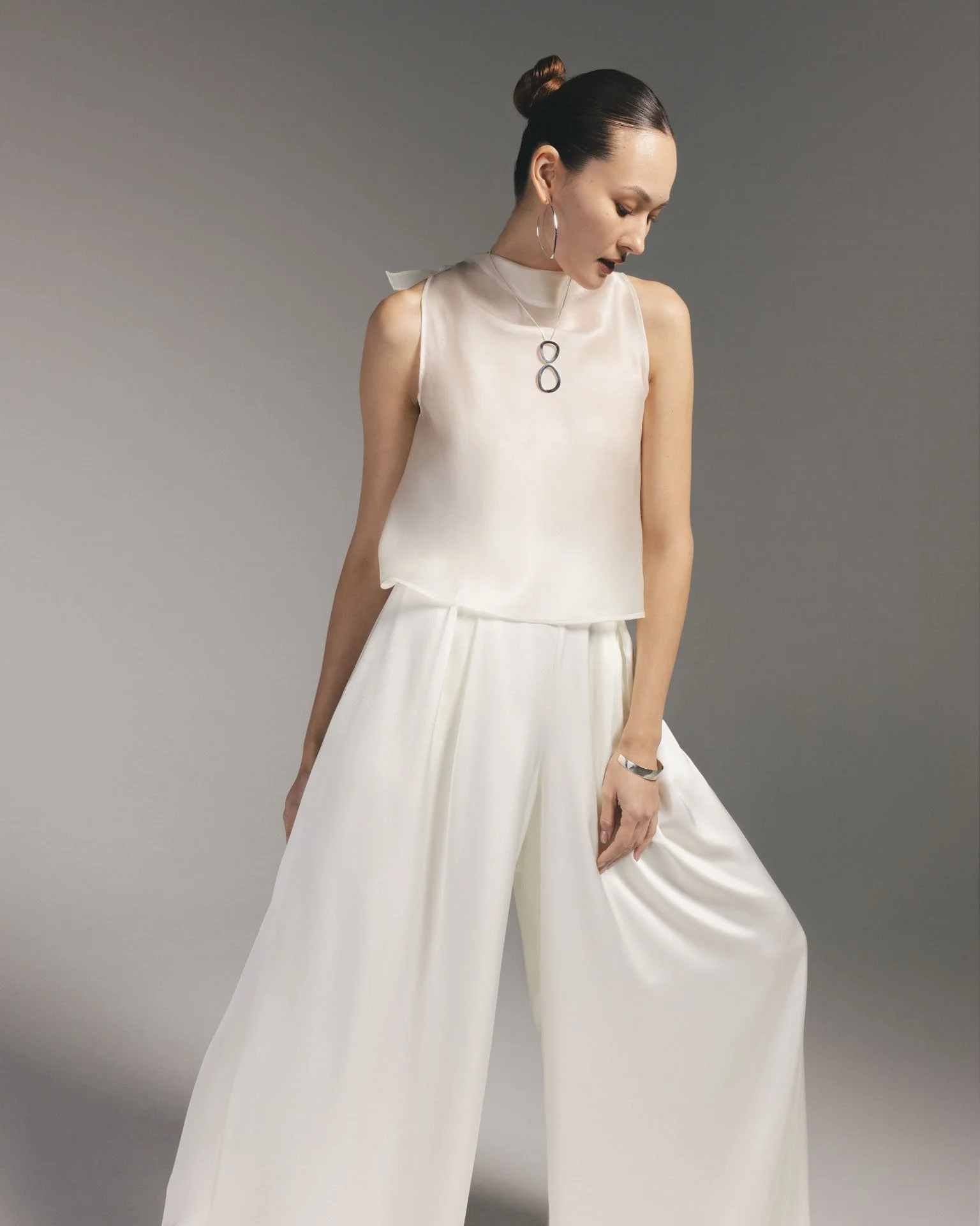 Model wearing a Karis white silk sleeveless mock-neck top paired with flowing white wide-leg trousers, accessorized with a silver pendant necklace and hoop earrings.