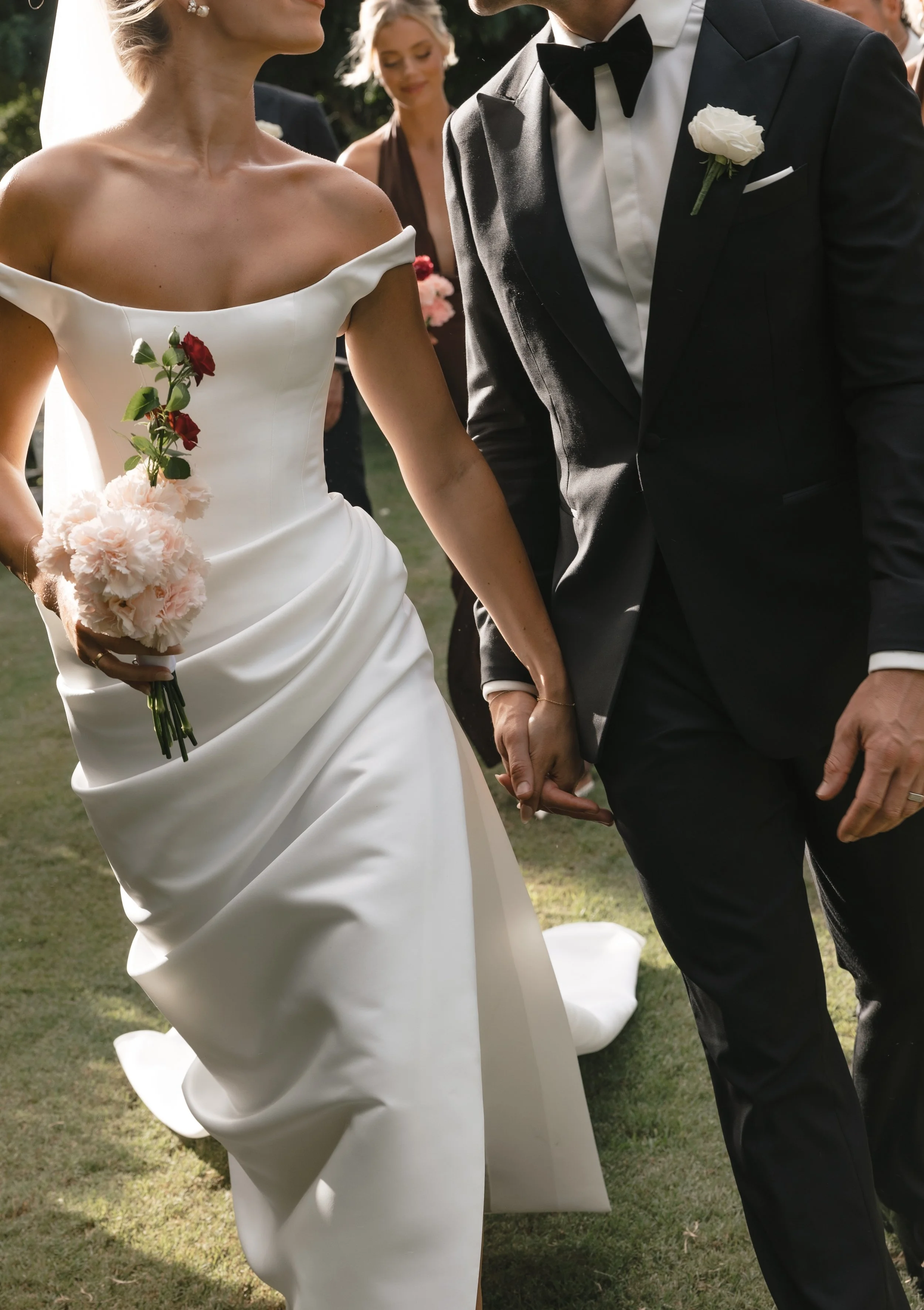 Close-up of a bride wearing a KYHA off-the-shoulder satin wedding dress featuring a clean minimalist neckline and an elegantly draped waist, holding a small floral bouquet while walking hand-in-hand with the groom.