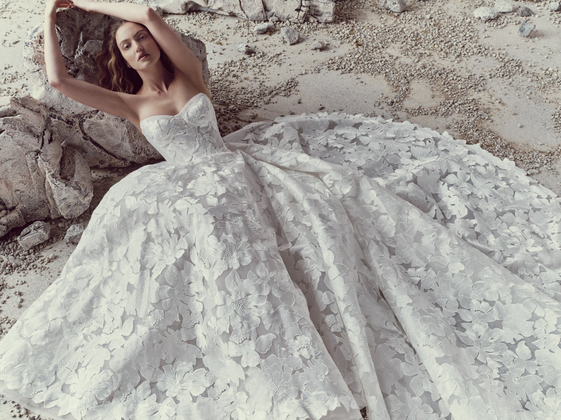 Model wearing a highly voluminous Rivini strapless ballgown wedding dress entirely covered in oversized 3D floral appliqués, leaning gracefully against a large rock in a sandy landscape.