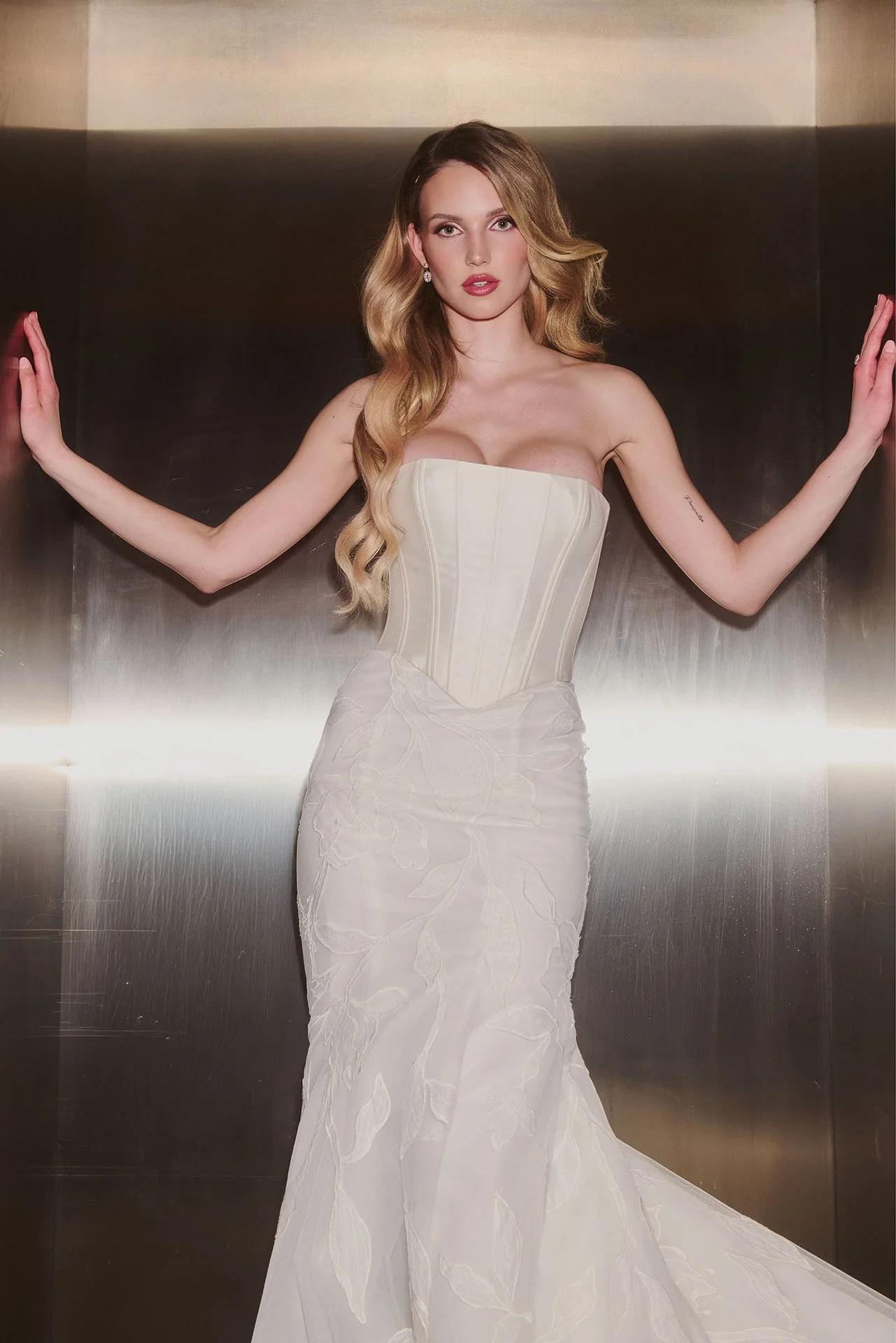Model wearing an Ines Di Santo strapless wedding dress featuring a minimalist, smooth satin corset bodice with a sharp basque waist, paired with a fitted skirt adorned with large tonal floral appliqués.