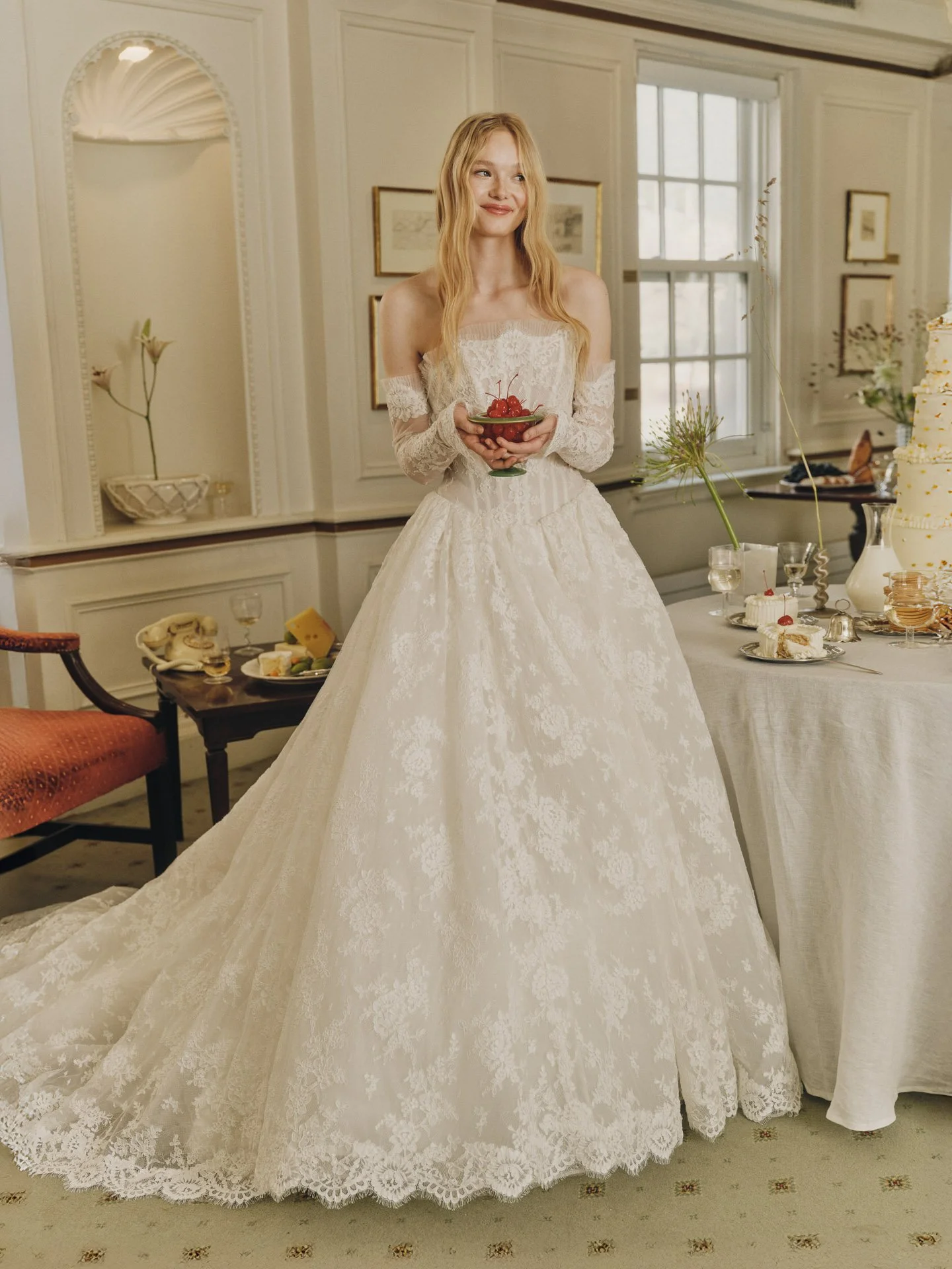 Model wearing an Alyne off-the-shoulder lace ballgown with separate sheer lace sleeves and a full train, holding a small bouquet in an elegant vintage-panelled room with a cake and telephone.
