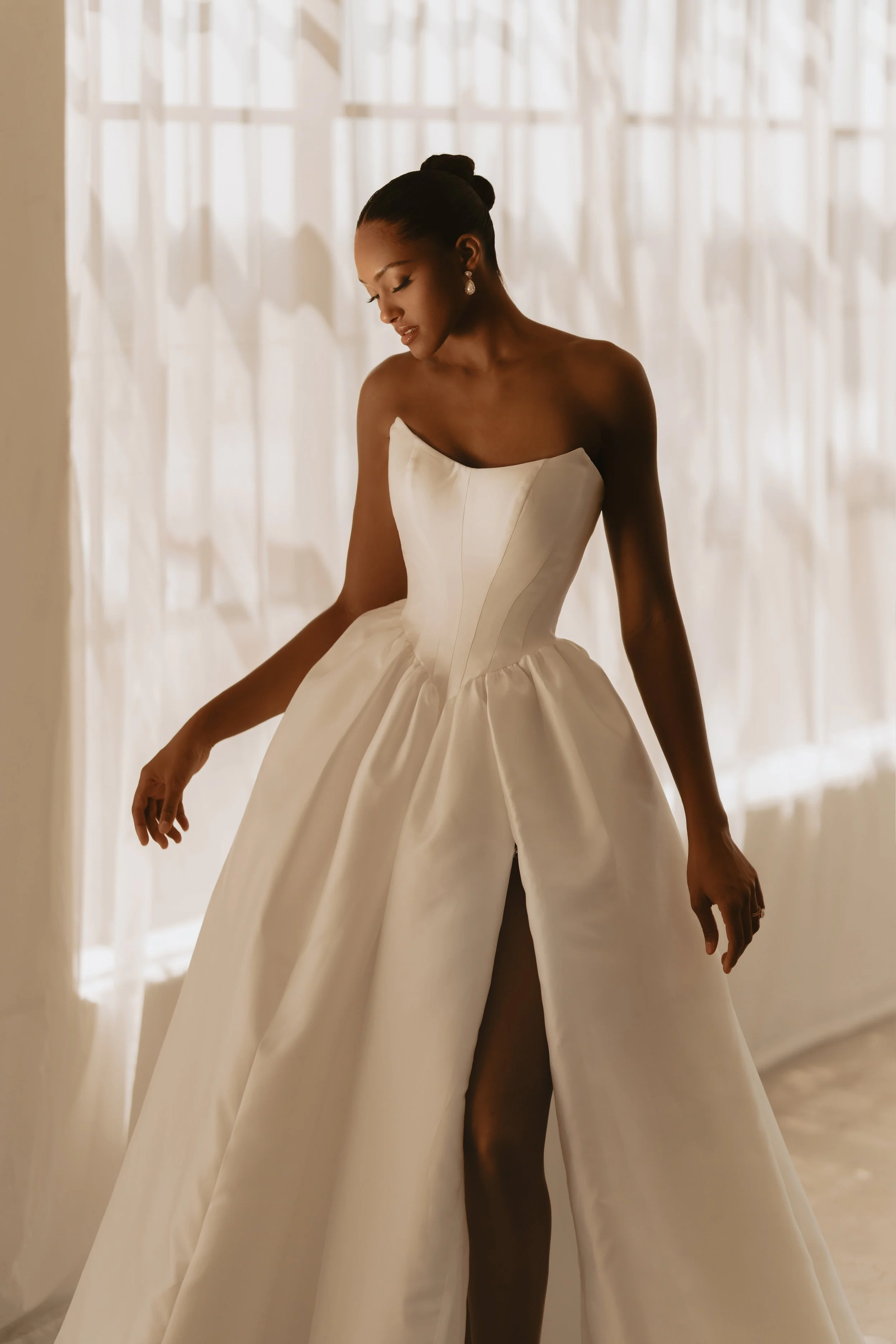 Model wearing a Martina Liana strapless satin ballgown wedding dress with a structured basque waist and a high leg slit, standing in soft, natural indoor lighting.