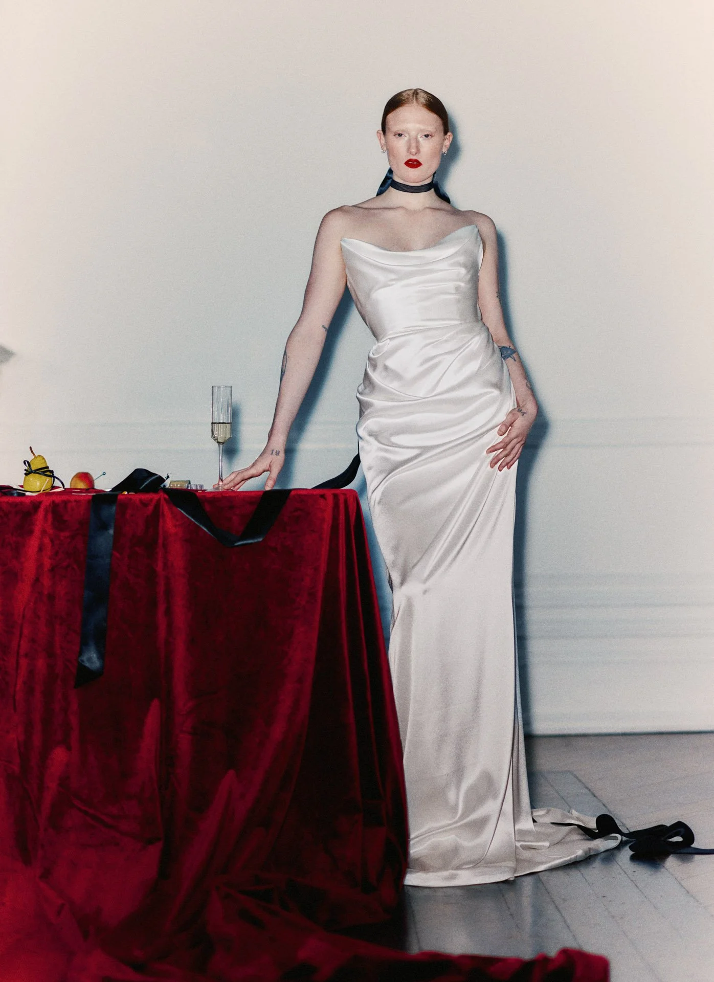 Model wearing a Former Girlfriend strapless satin wedding dress with an elegantly draped and ruched bodice, styled with a black ribbon choker and standing next to a red velvet-draped table