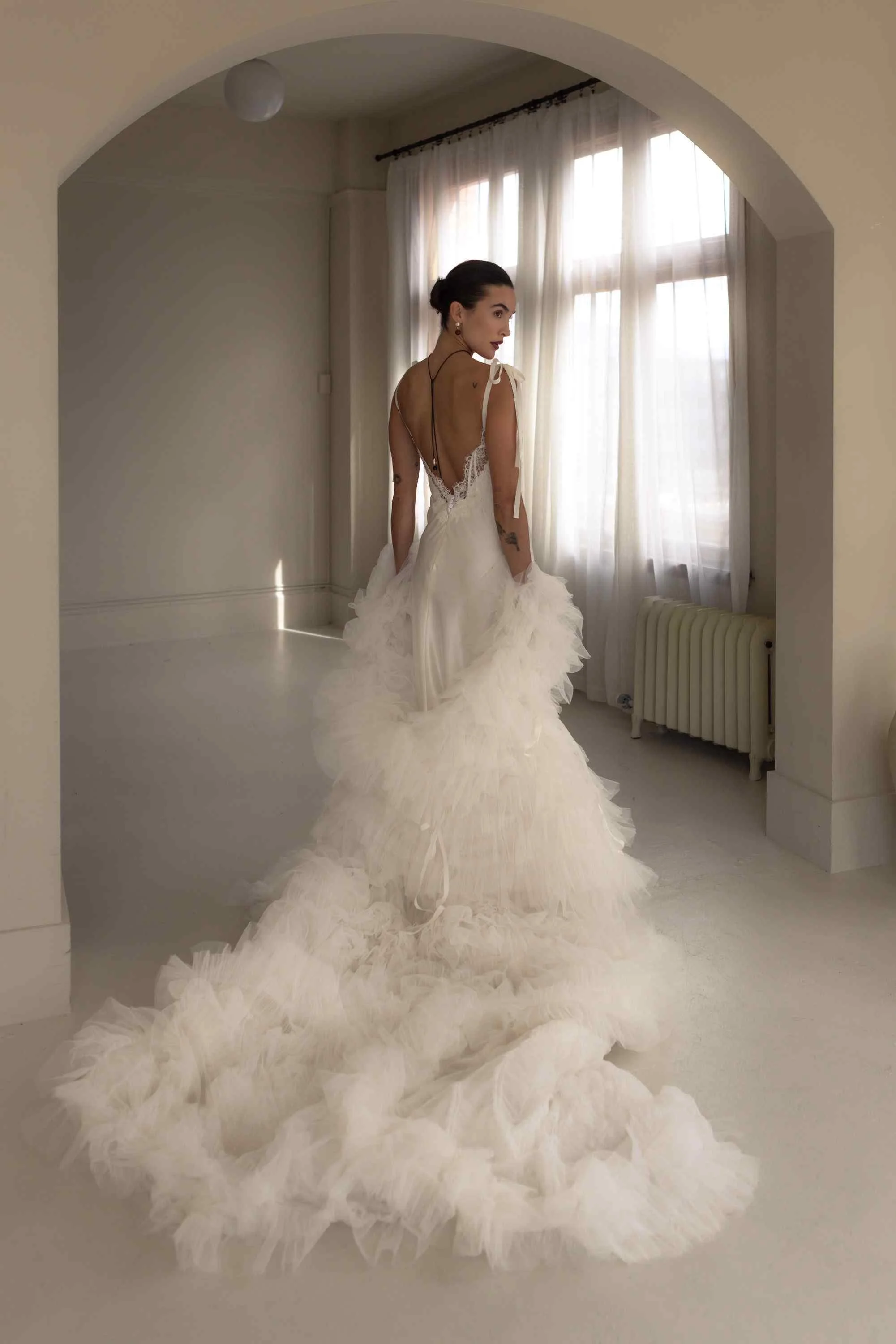 Back view of a model wearing an OUMA slip wedding dress featuring a low open back with delicate lace trim, styled with a massive, dramatic ruffled tulle bridal cape trailing across the floor.